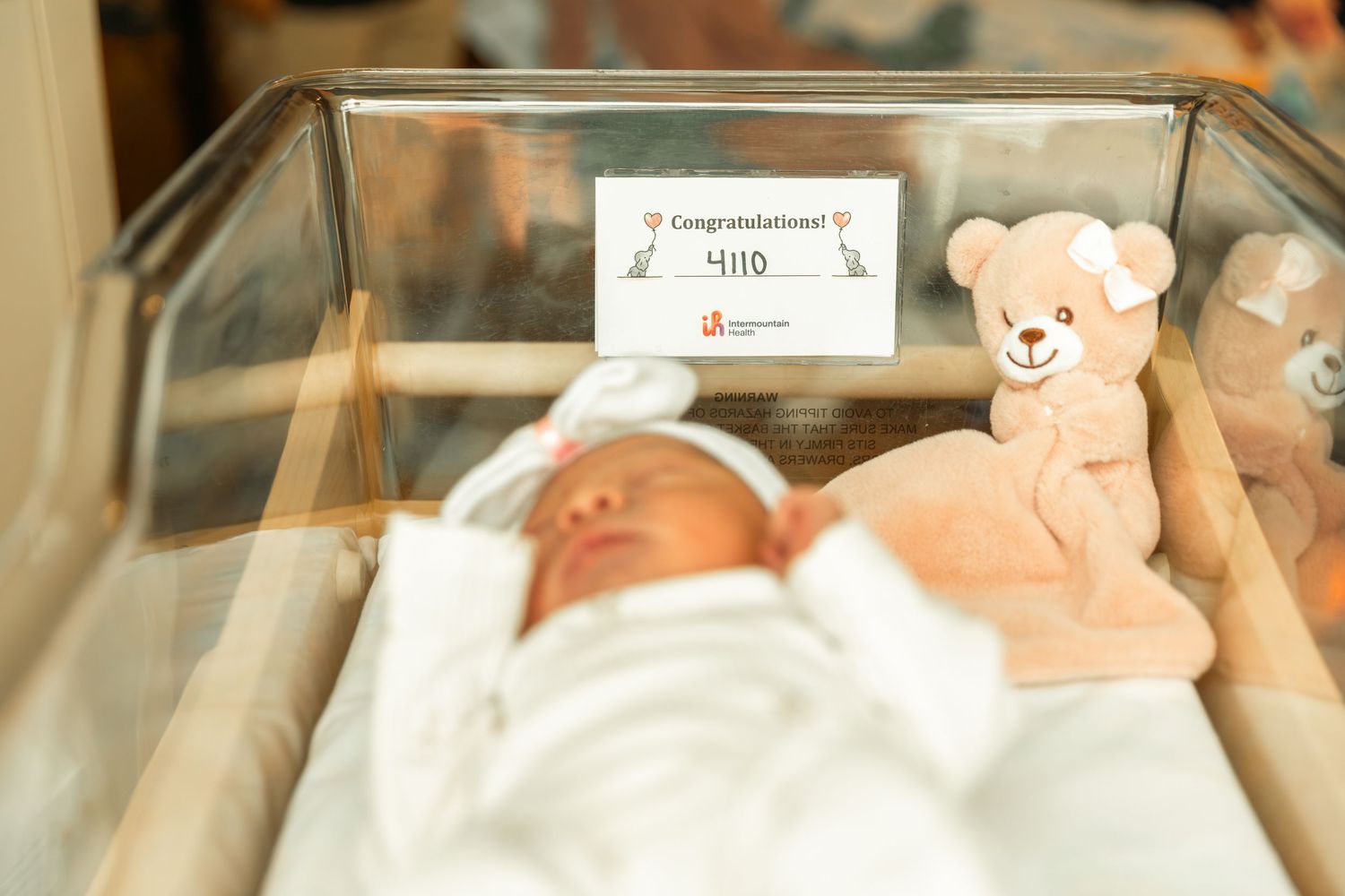Newborn baby sleeping in hospital bassinet with a congratulations sign and teddy bear toy.