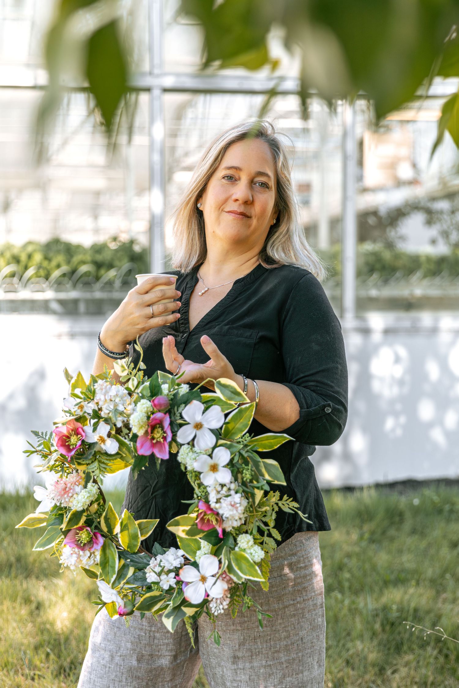 Woman in black top holding a floral wreath with pink and white flowers outdoors near a greenhouse.