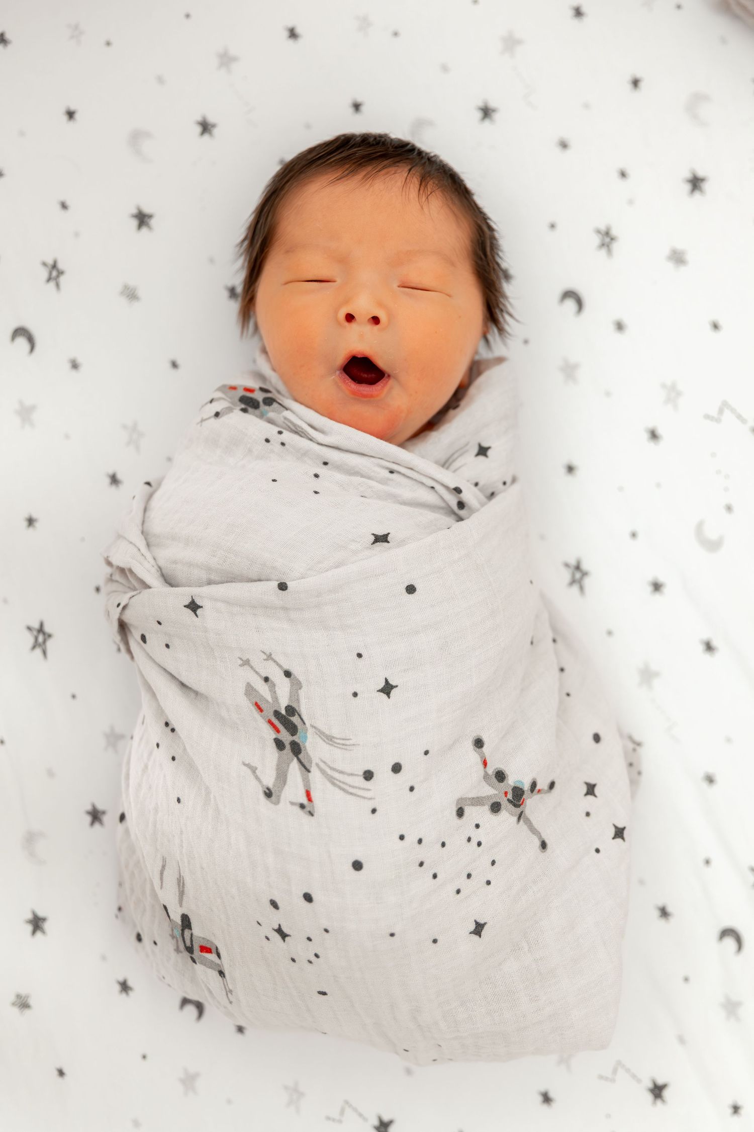 Newborn baby swaddled in star-print blanket yawning, lying on celestial patterned white sheet.