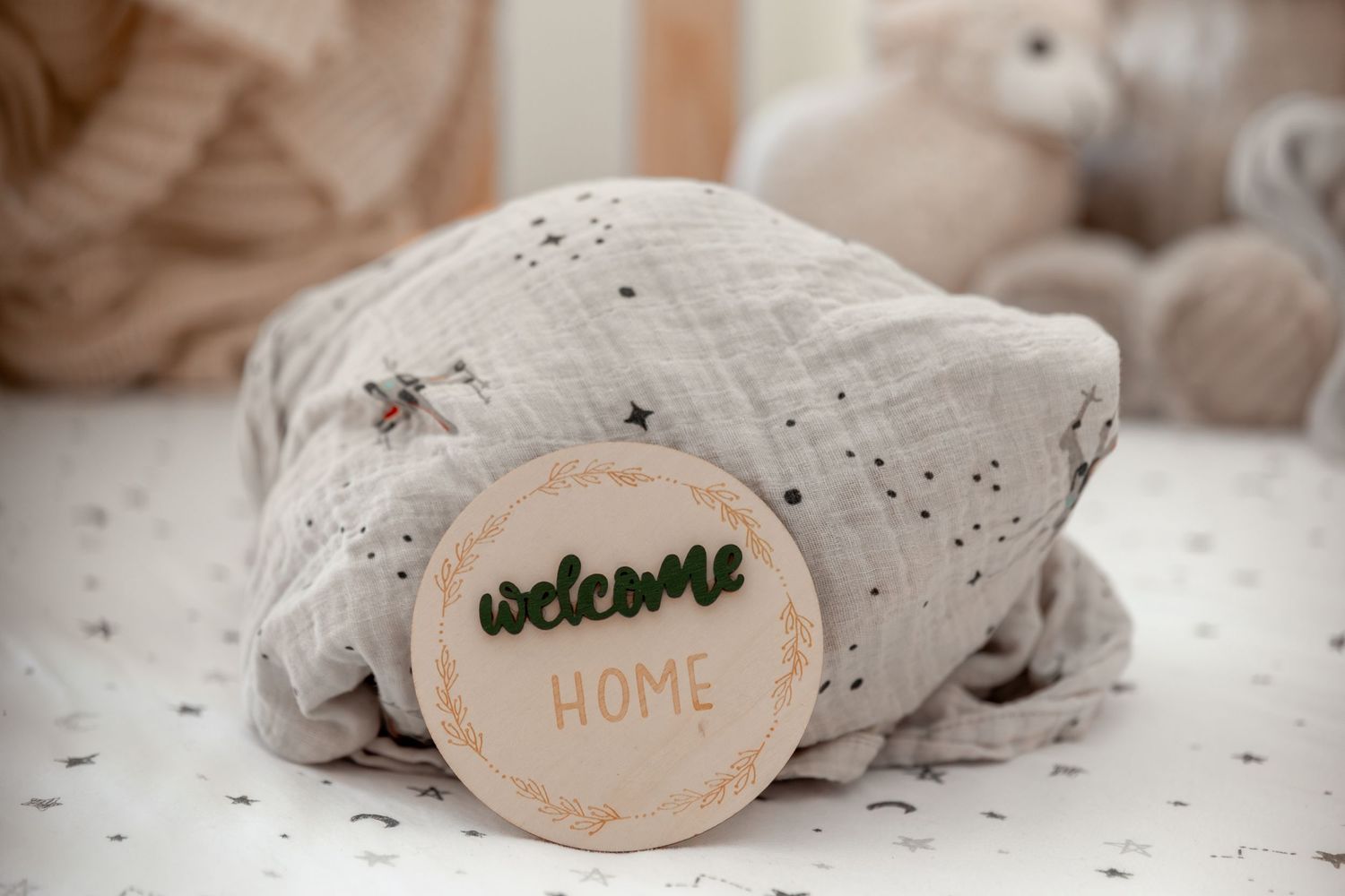 A wooden welcome home sign rests against a soft gray muslin blanket with star print in a cozy nursery setting.