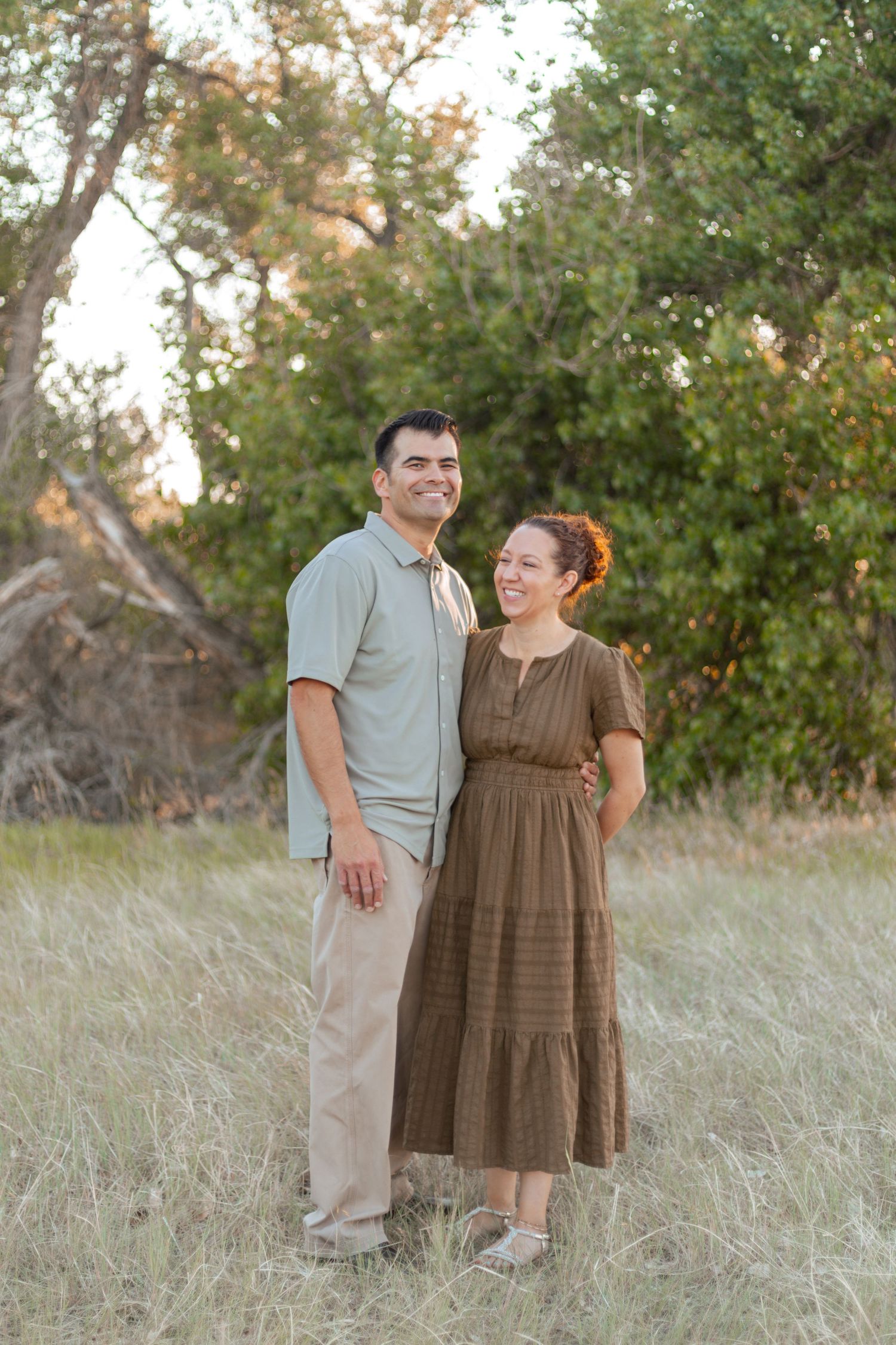 A smiling couple stands together in a grassy field with green trees in the background during golden hour.