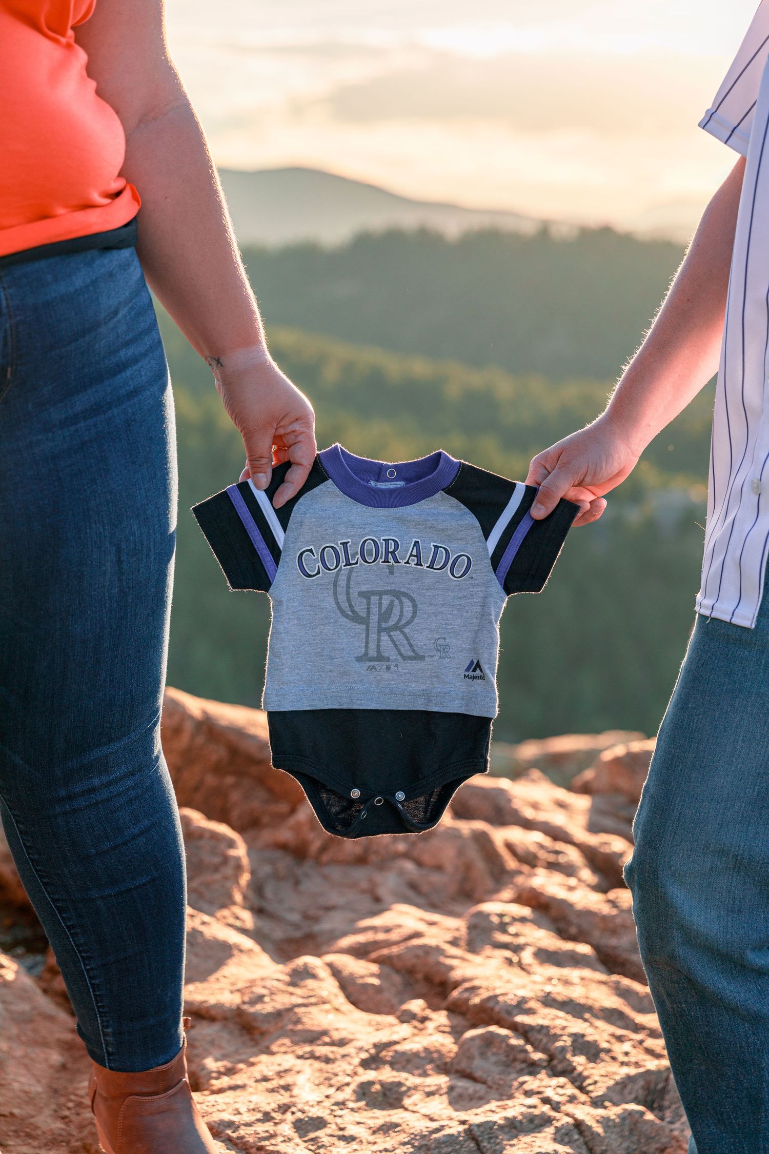 Small Colorado Rockies baby onesie held up against mountain sunset backdrop.