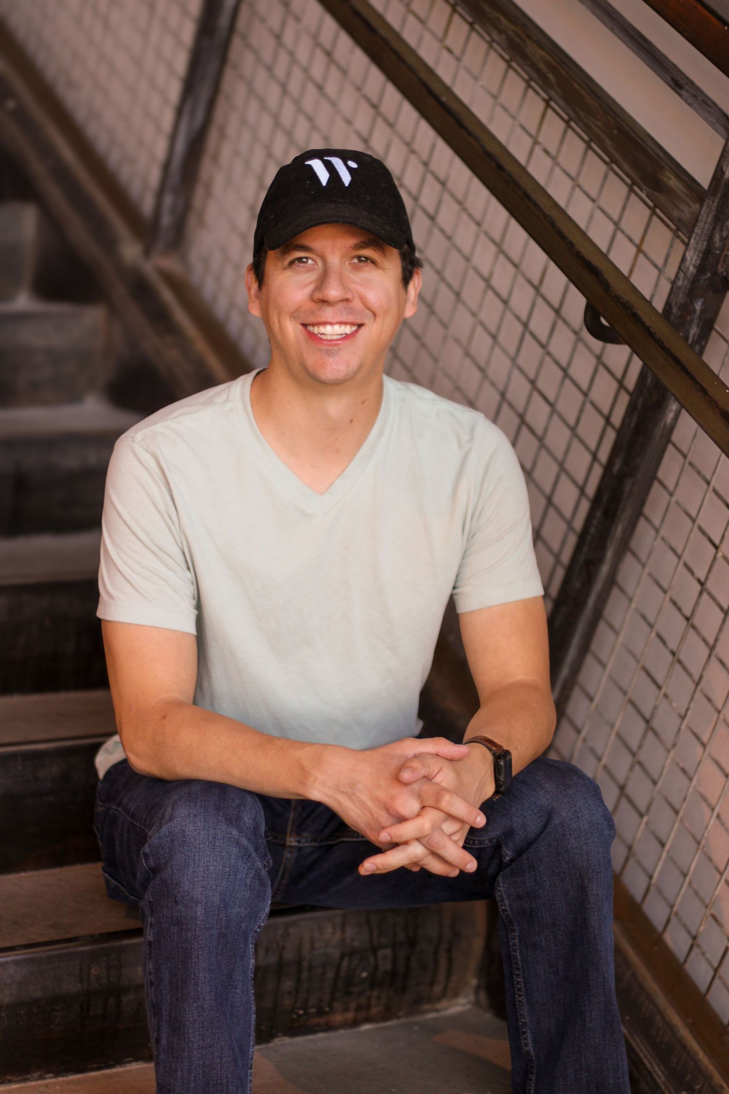 A person in a white v-neck t-shirt and baseball cap sits casually on wooden stairs.
