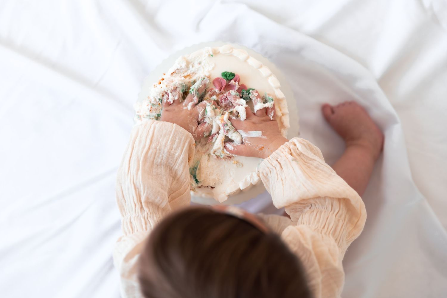 Overhead view of a baby in a peach outfit smashing a decorated white cake on a white sheet.