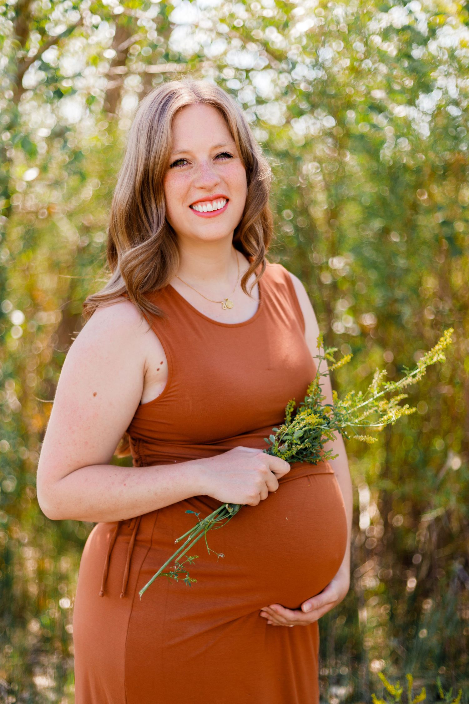 Woman in rust-colored dress holding greenery outdoors in natural setting.
