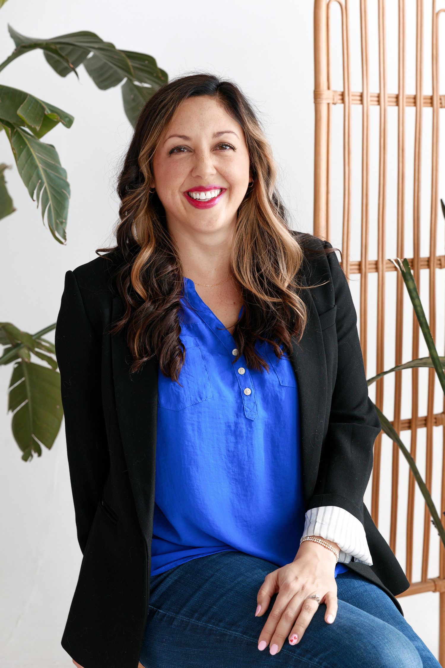 Professional portrait of a woman in a blue top and black blazer smiling against a white wall with tropical plants.