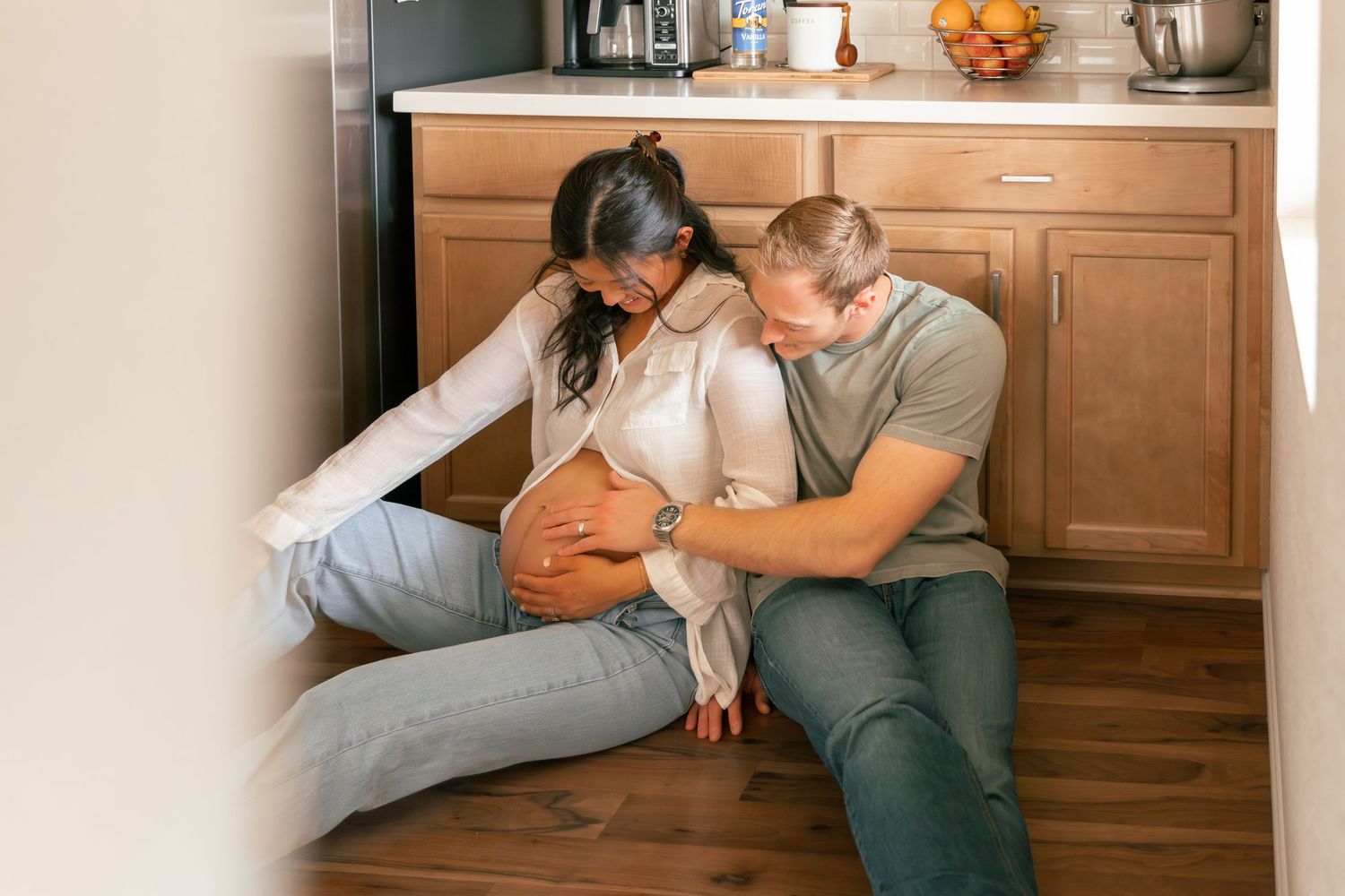 A couple sits together on the floor of their home with their orange cat, sharing a tender moment.