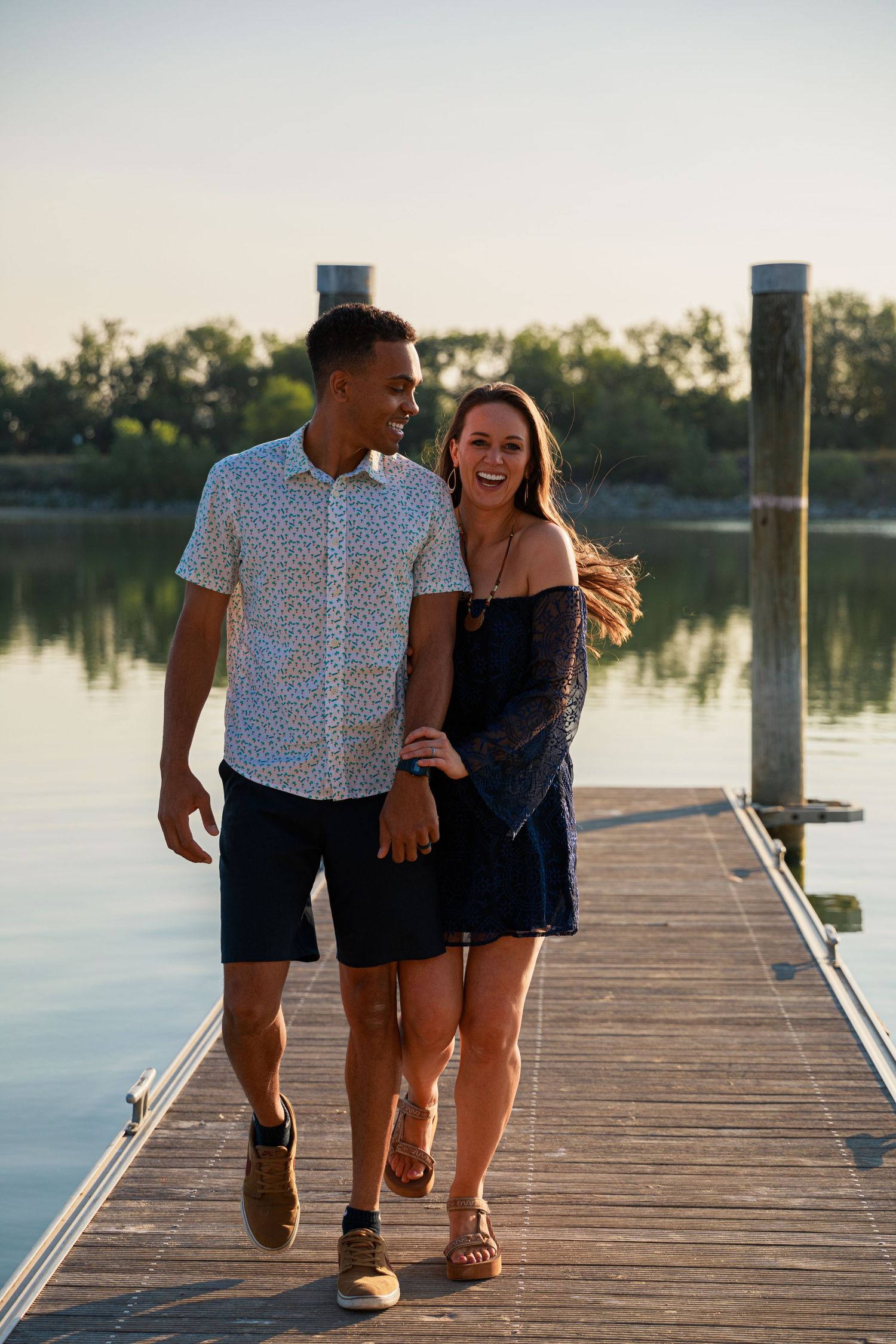 A couple walks and laughs together on a wooden dock by a serene lake at sunset.