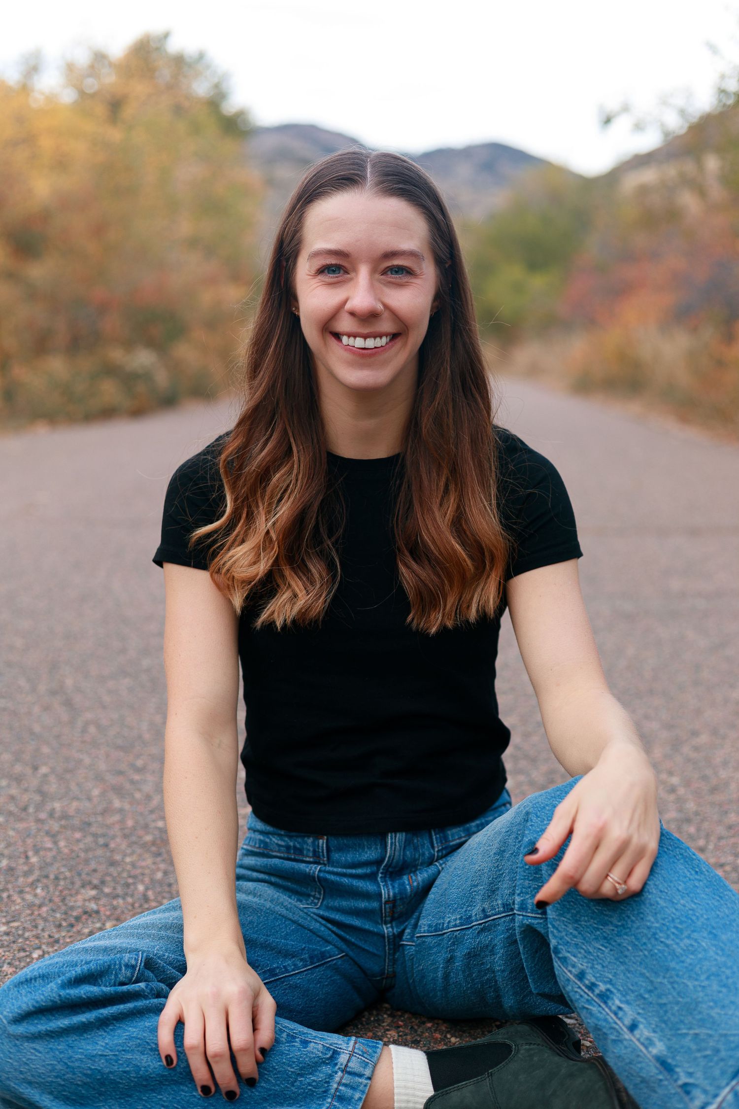 Young woman with long brown hair smiling, sitting on a road in jeans and black tee, with autumn trees and mountains behind.