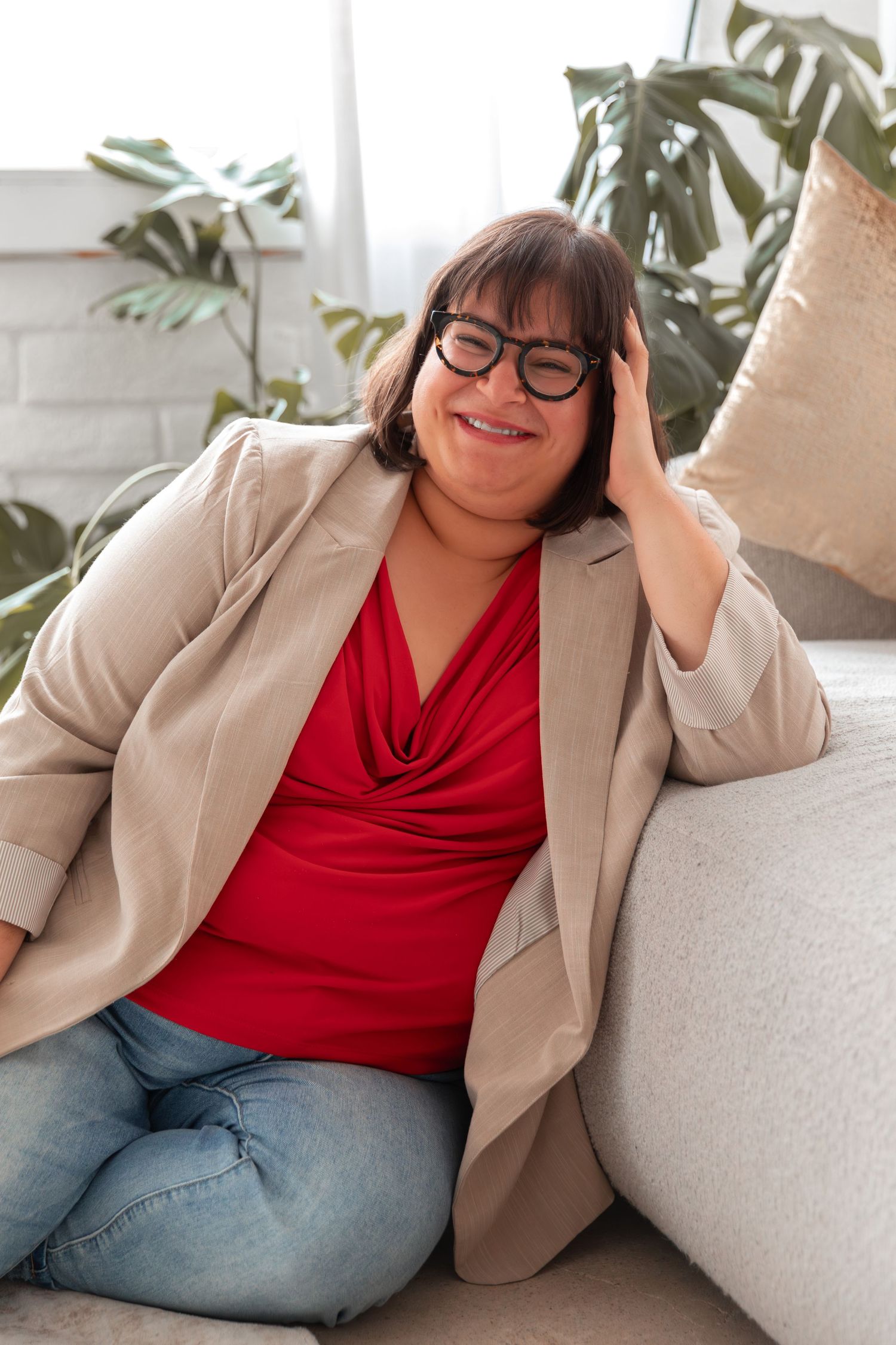 Plus-size woman in red top, blazer, and glasses smiling while sitting on a couch indoors.