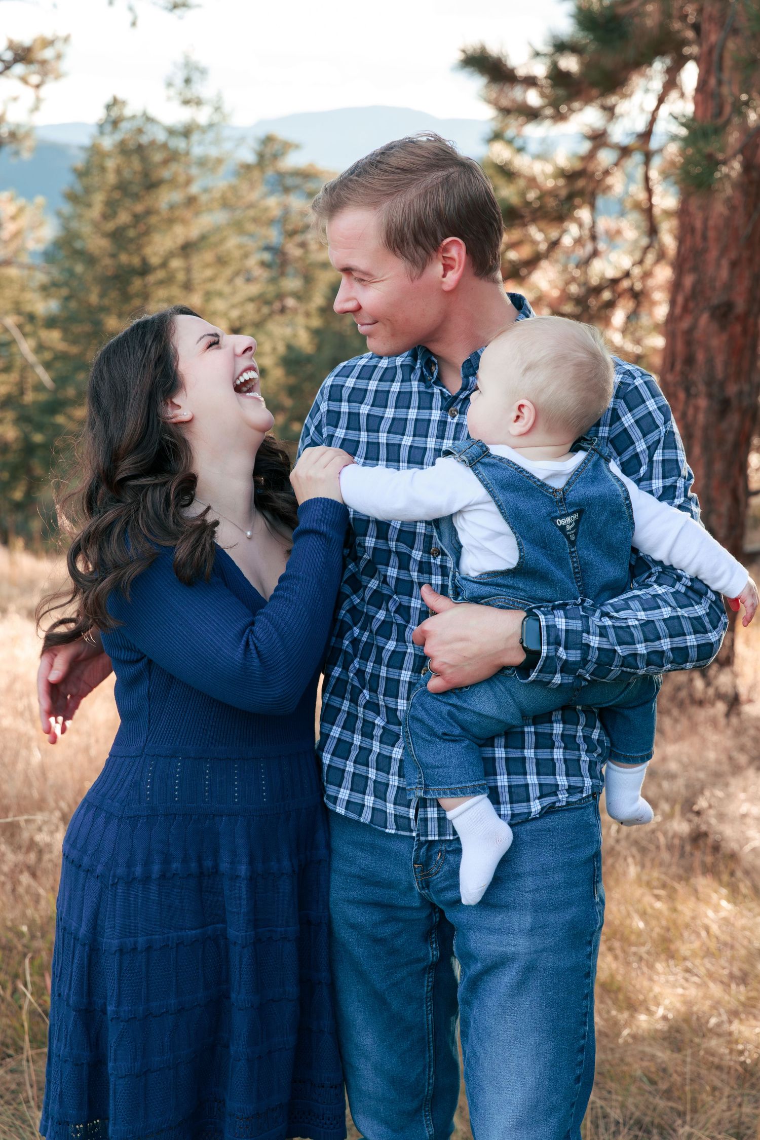 Happy young family with baby outdoors in pine forest, laughing couple holding infant in blue outfits.