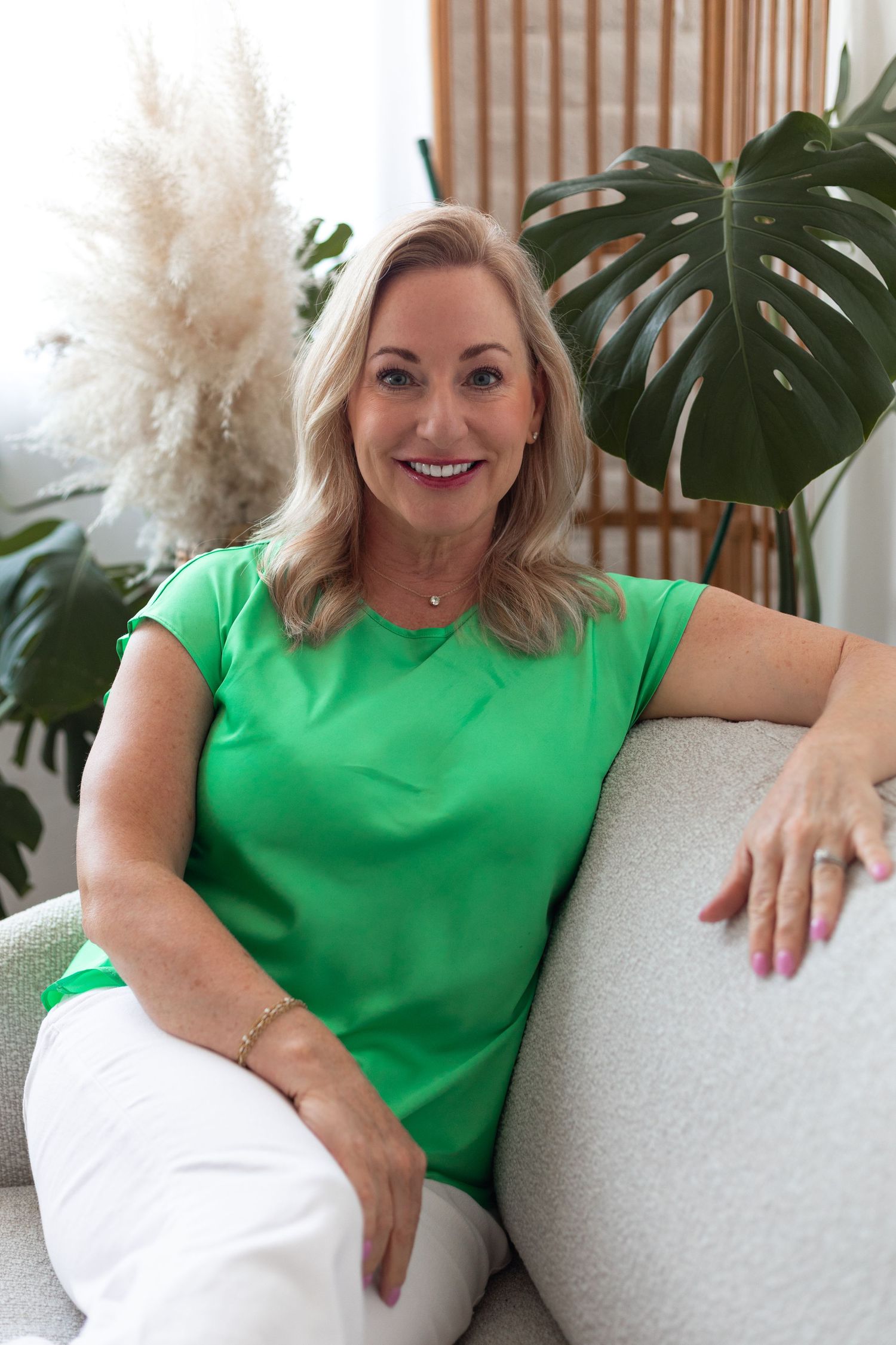 A woman in a bright green top poses casually on a white couch with tropical plants in background.