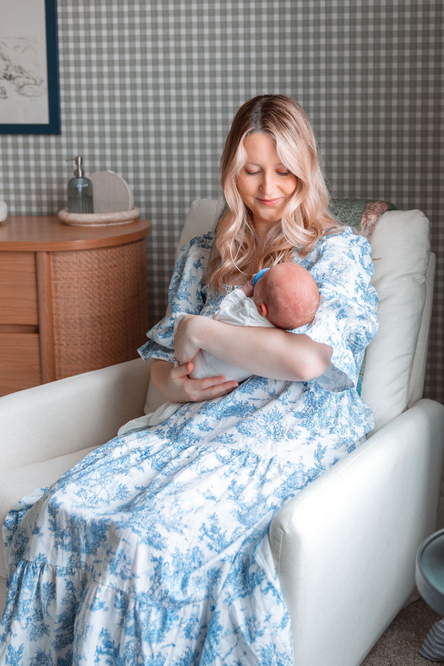 Nursery scene with white glider chair and blue floral dress against gingham wallpaper.