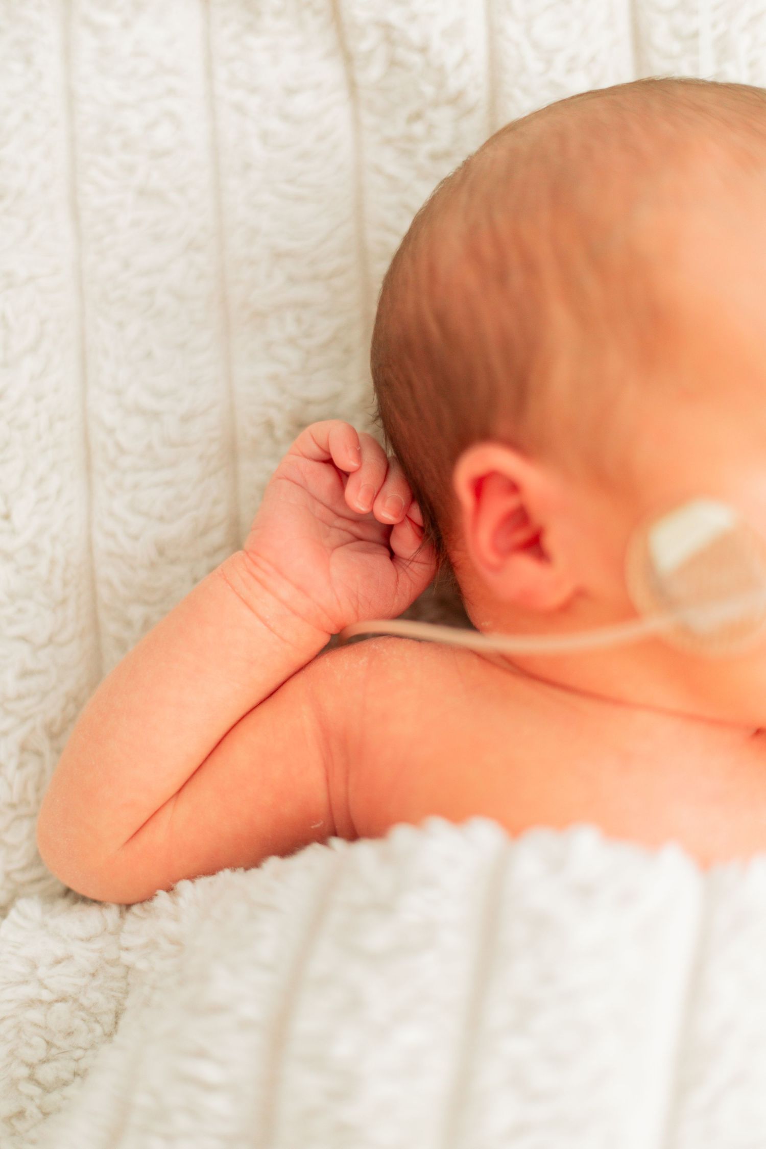 Close-up of a sleeping newborn baby lying on a white knit blanket, with a small oxygen tube near their ear.