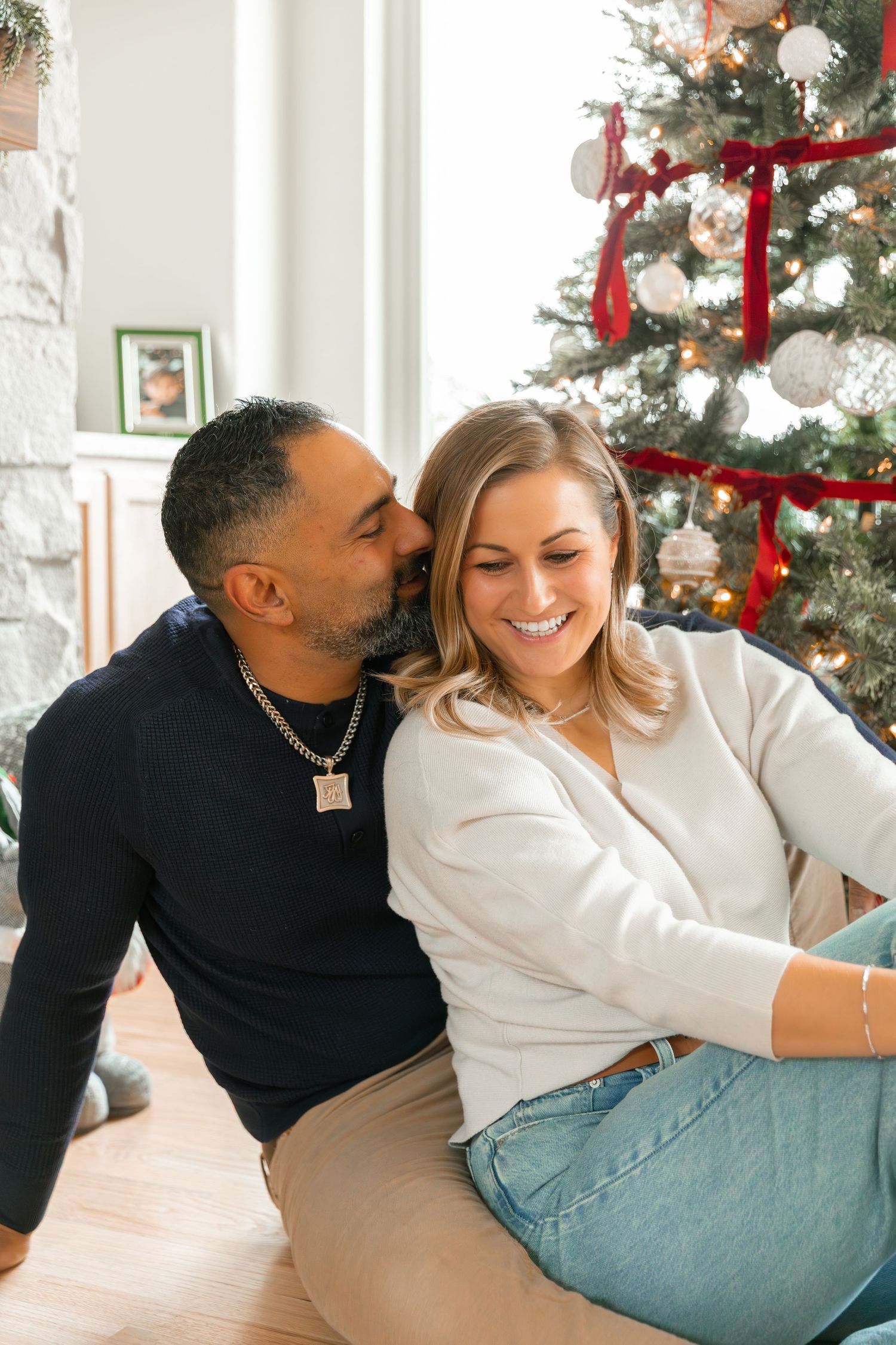 A couple shares a romantic kiss near a decorated Christmas tree, with the woman smiling joyfully in a cozy holiday setting.