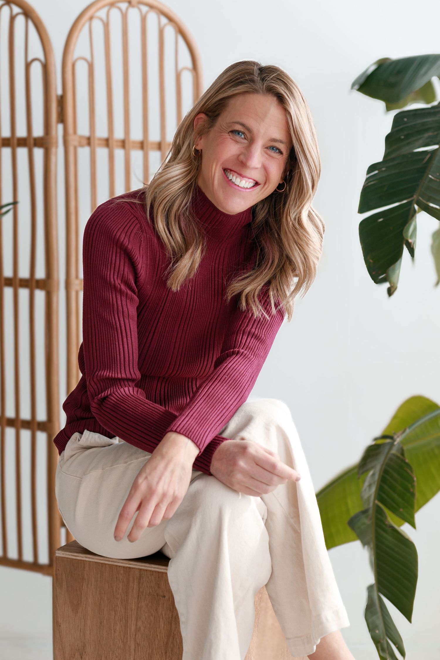 Woman in burgundy sweater and white pants laughing while sitting on wooden furniture.