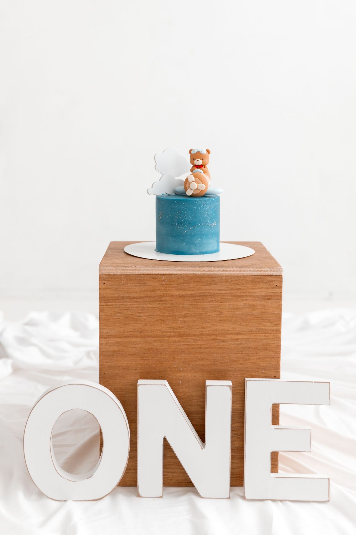 Blue first birthday cake with teddy bear topper on wooden block, surrounded by white ONE letters.