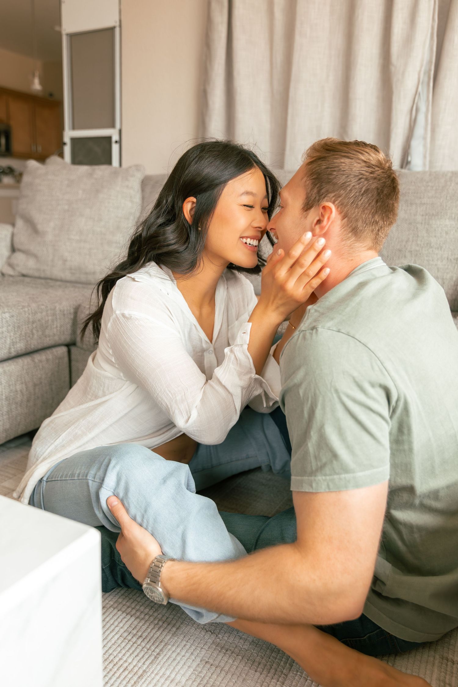 A couple shares intimate kisses while relaxing together on a couch in a bright, cozy living room setting.