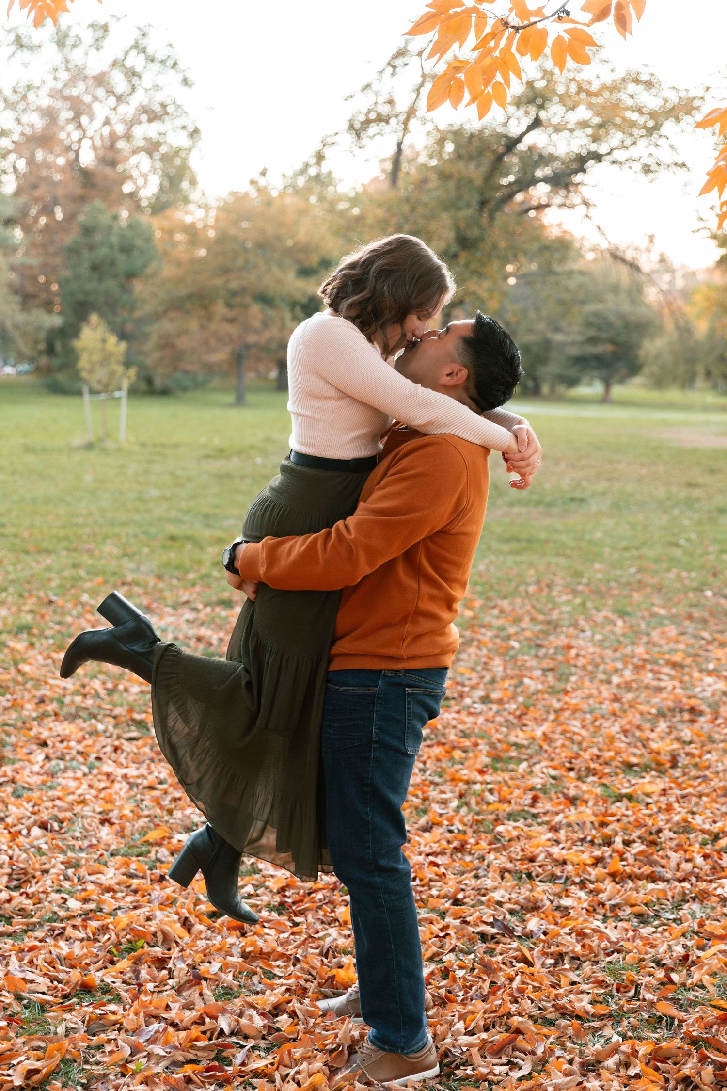Two people playfully embrace in a park filled with vibrant autumn leaves.