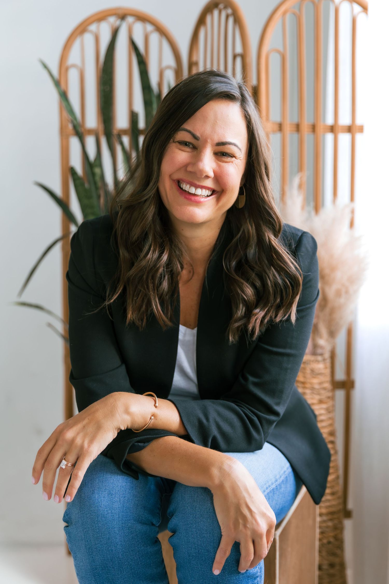 Professional portrait of woman in blazer and jeans sitting casually against neutral background.