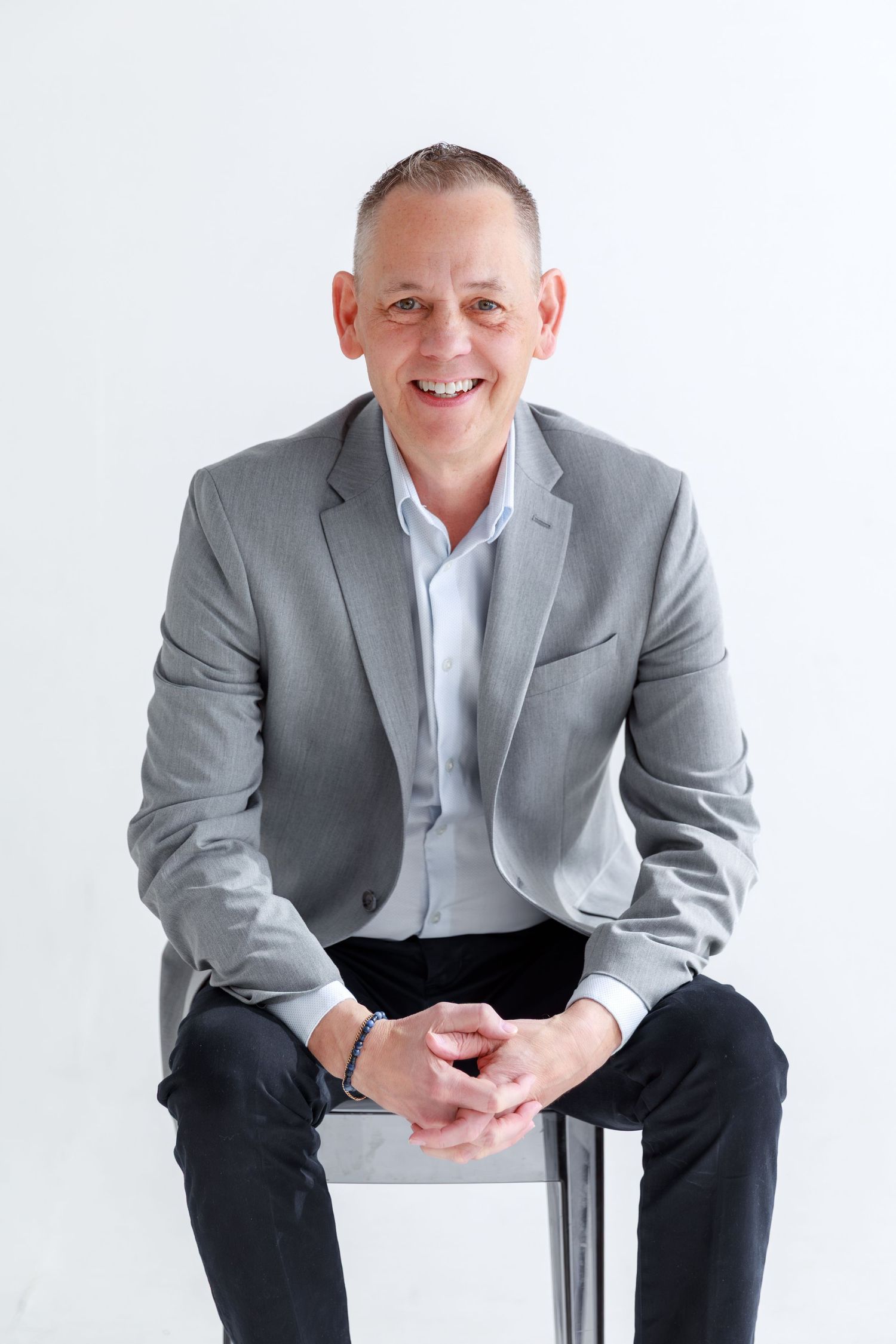Professional studio portrait of someone in a gray suit jacket and light blue shirt sitting casually on a white stool.