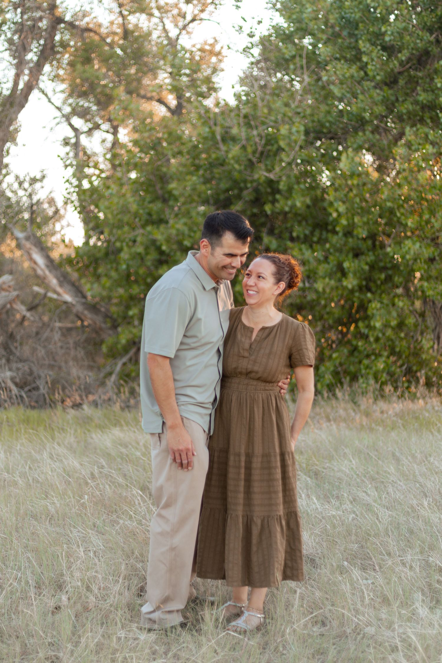 Couple smiling and embracing in a golden grass field with green trees, wearing earth-tone casual outfits.