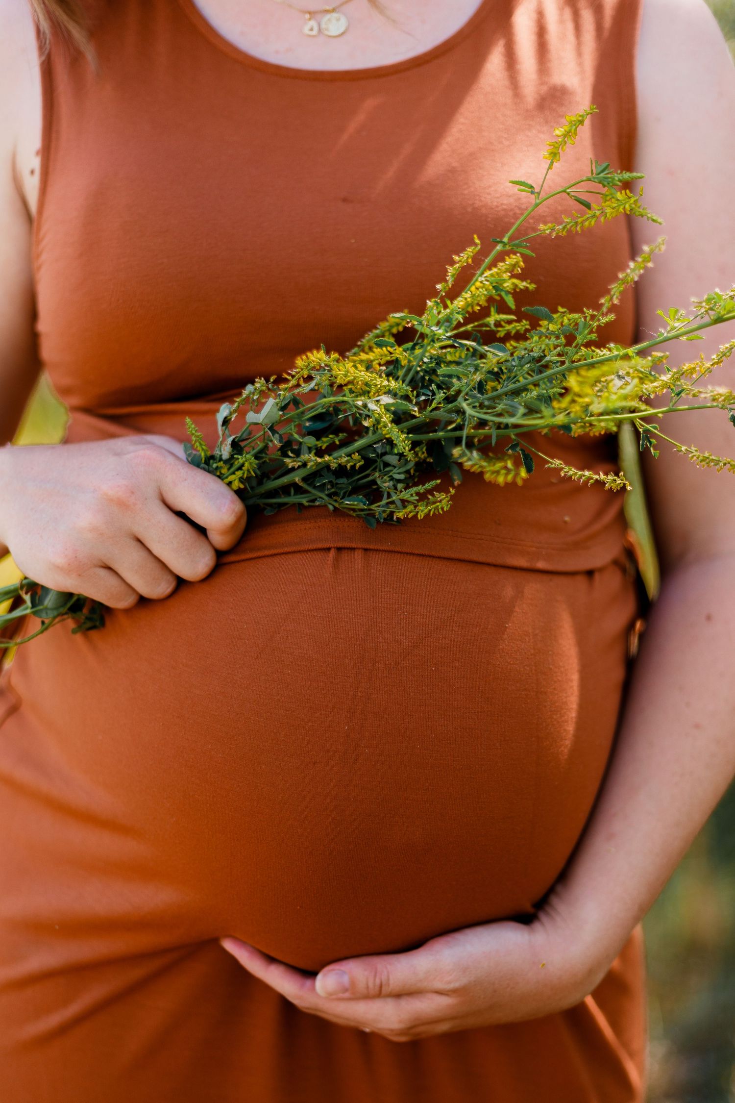 A pregnant person in a rust-colored dress holds wildflowers against their belly.