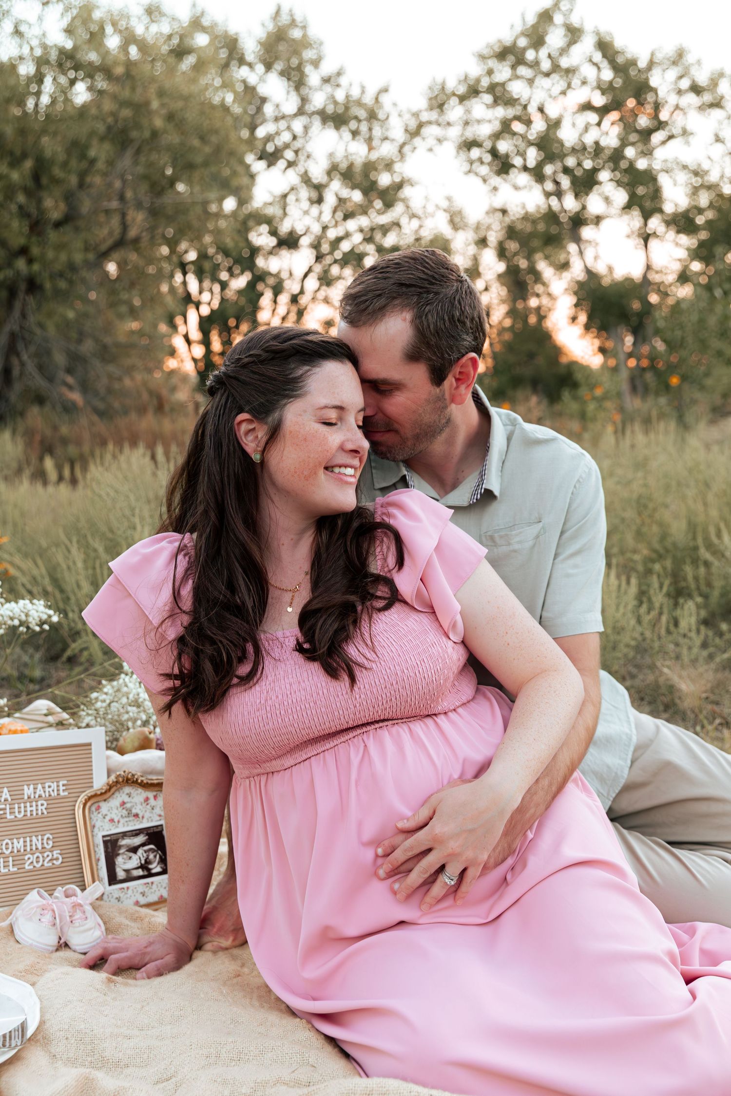 A romantic maternity photoshoot in a field at sunset shows a couple sharing tender moments on a blanket.