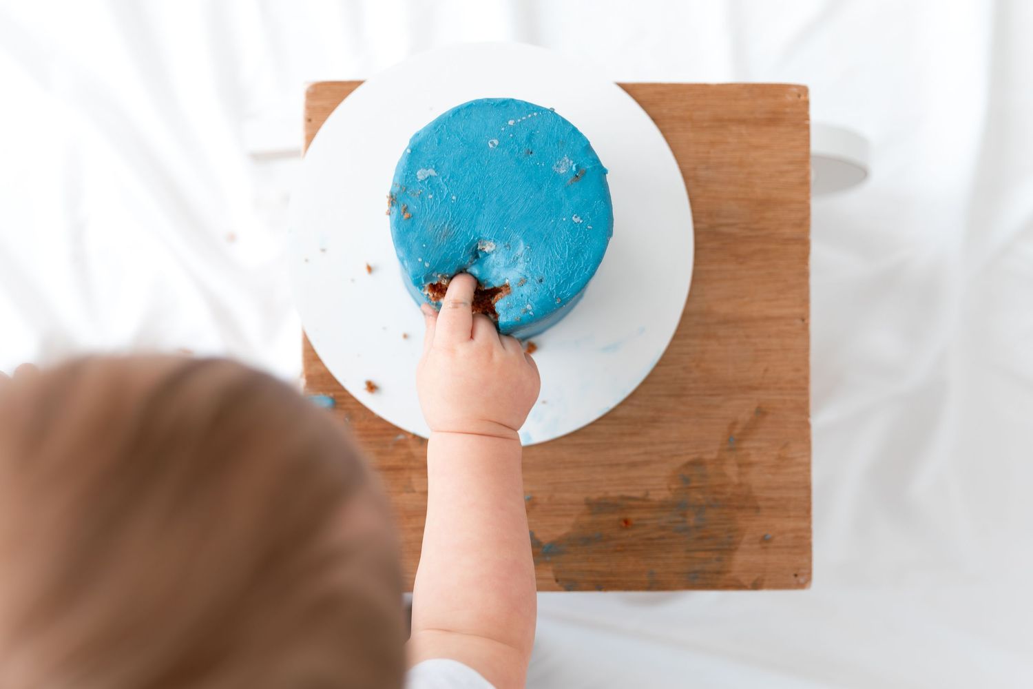 Baby reaching into a blue frosted birthday smash cake on a white plate and wooden cutting board.