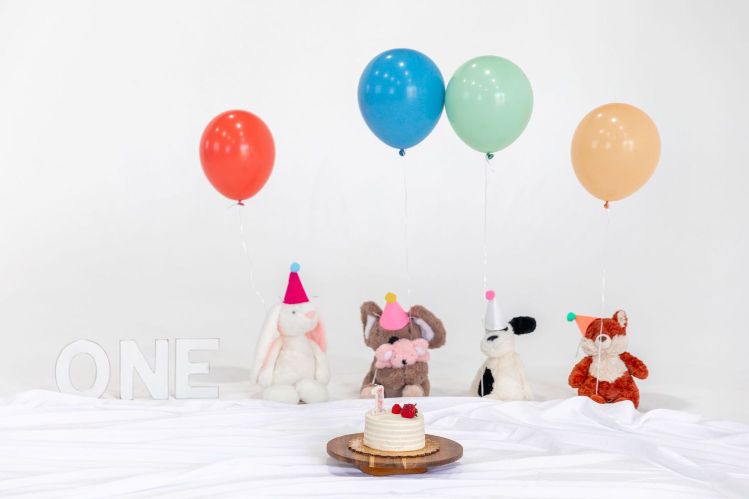 Stuffed animal toys with party hats sit around a small birthday cake with colorful balloons and ONE sign.