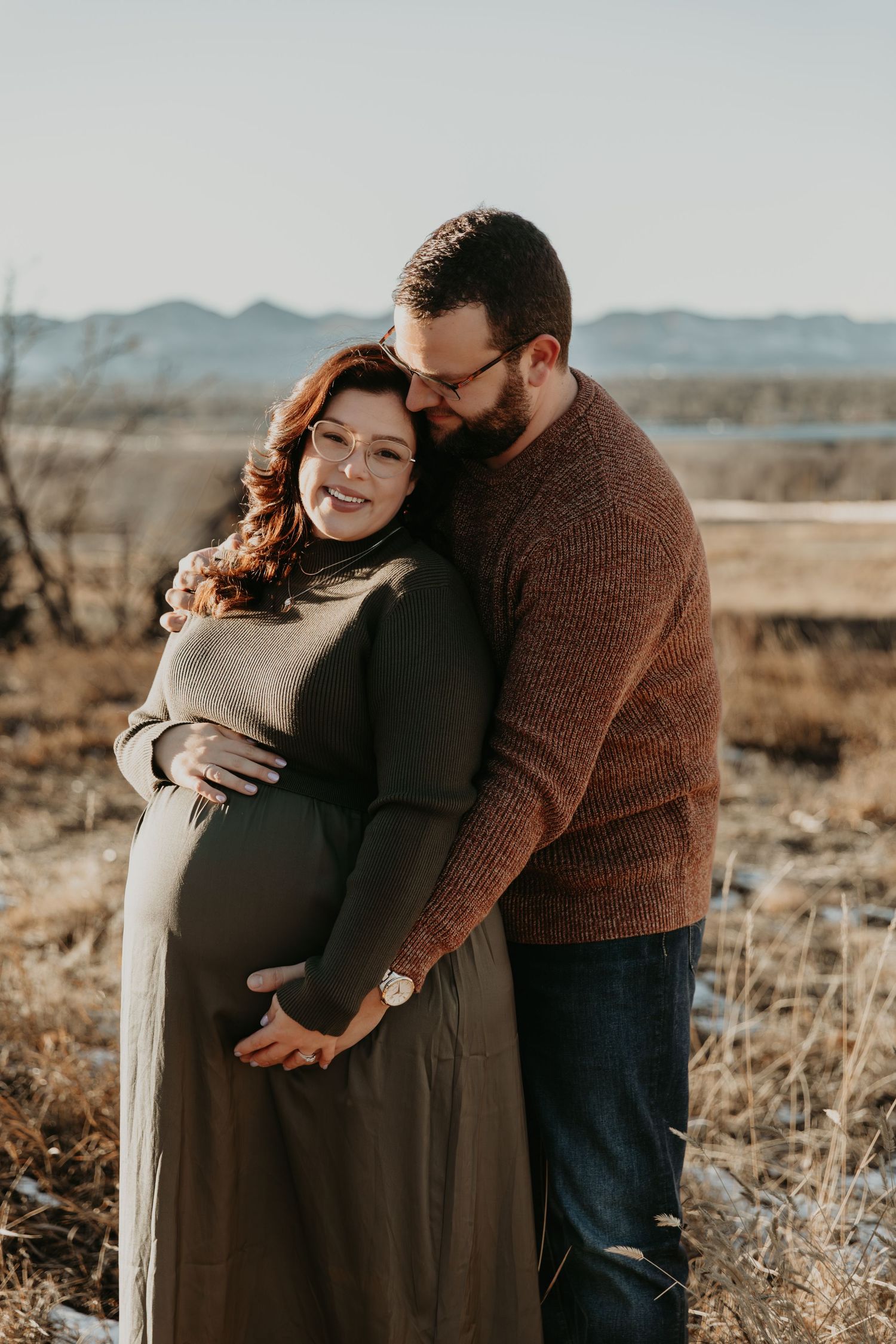 Couple shares intimate moments in a winter field during a maternity photoshoot with mountains in the background.
