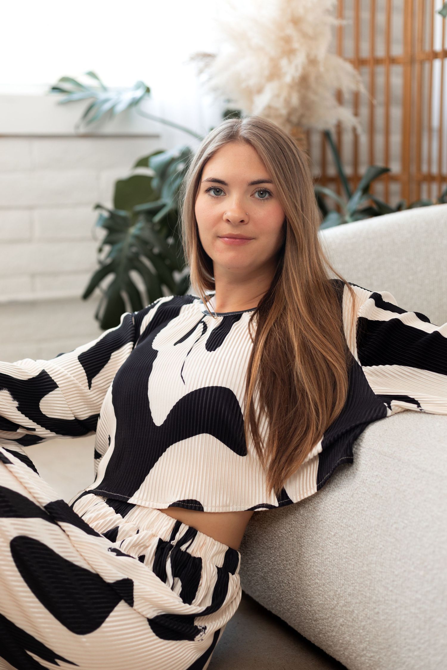 A woman relaxes on a couch wearing a black and white patterned dress near indoor plants.