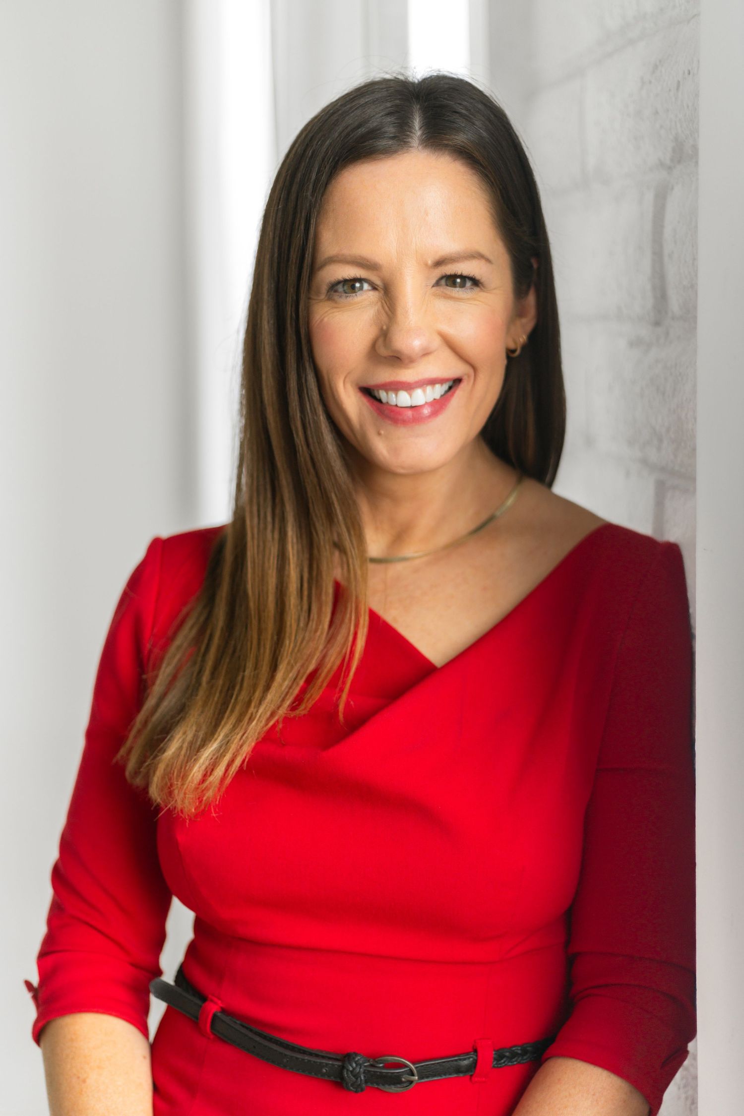 Smiling woman with long brown hair wearing a red dress and black belt, posing confidently in a bright modern setting.