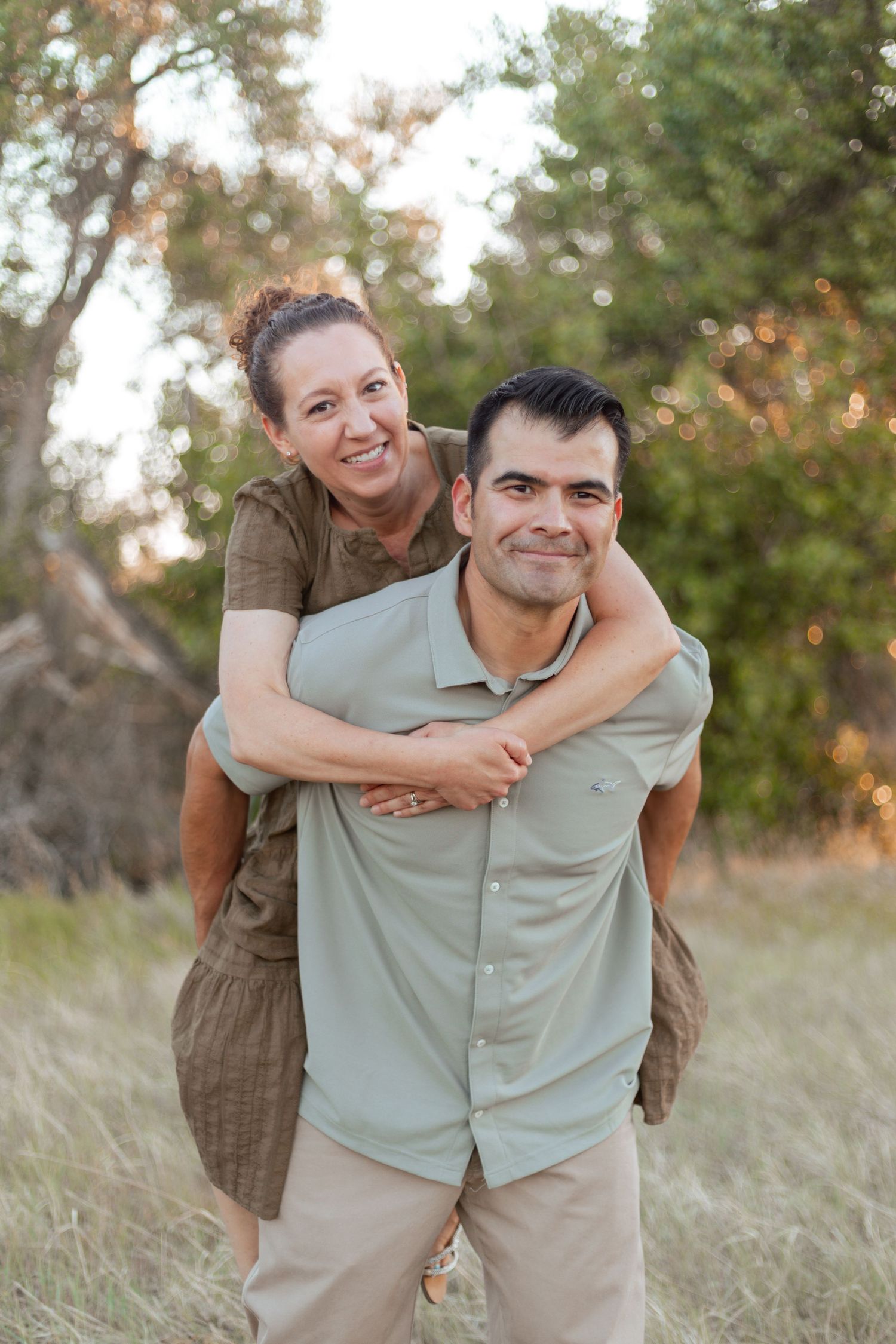 A woman piggybacks on a smiling man outdoors in a grassy field with trees in the background.