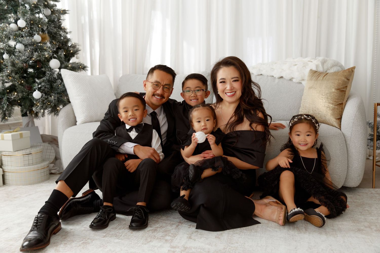Family of six in matching black outfits posing together near a Christmas tree in a bright, elegant studio setting.