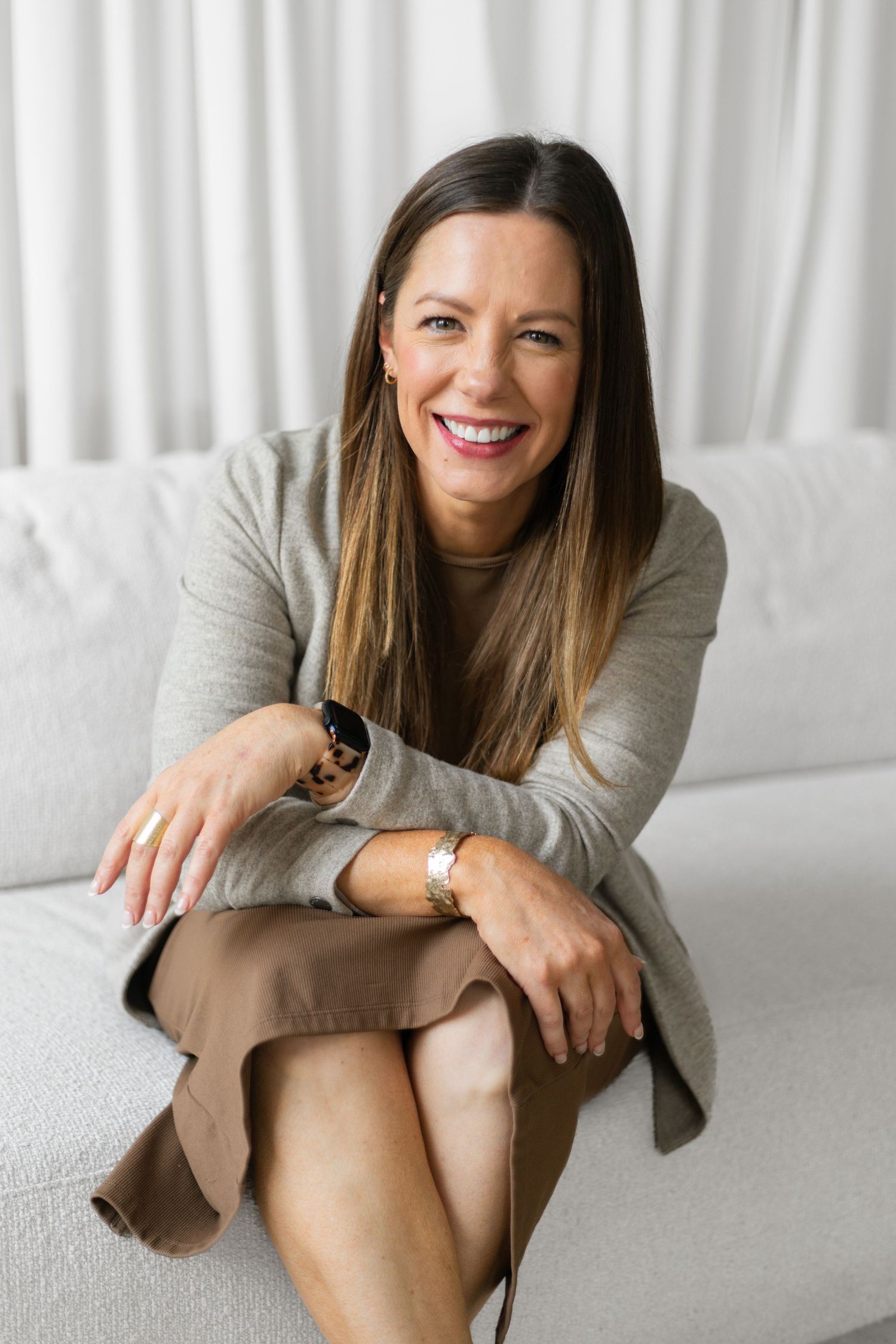 Smiling brunette woman in gray sweater and tan skirt sitting relaxed on white sofa in bright room.