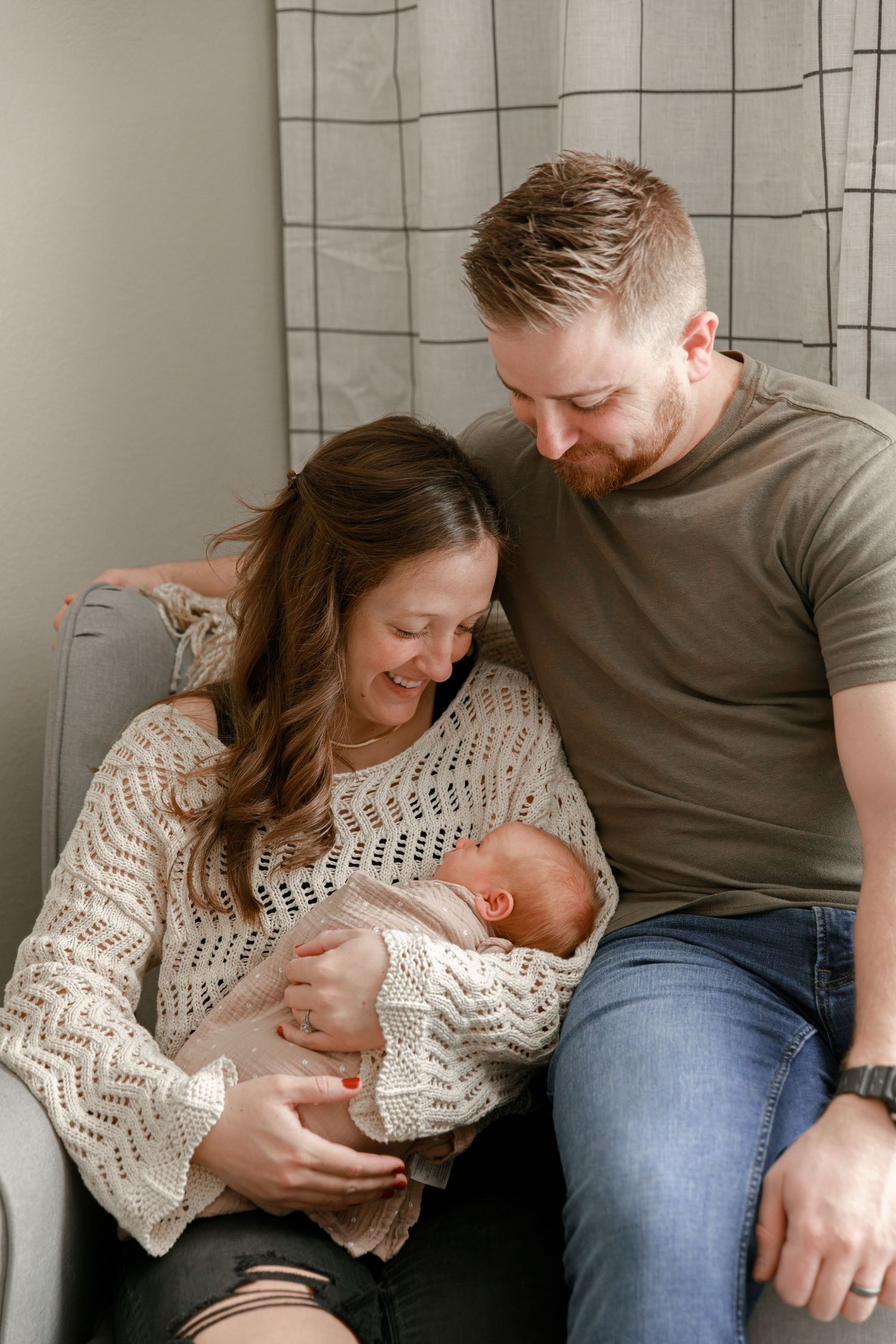 A couple sits on a gray couch sharing an intimate moment with their newborn baby in a cozy living room setting.