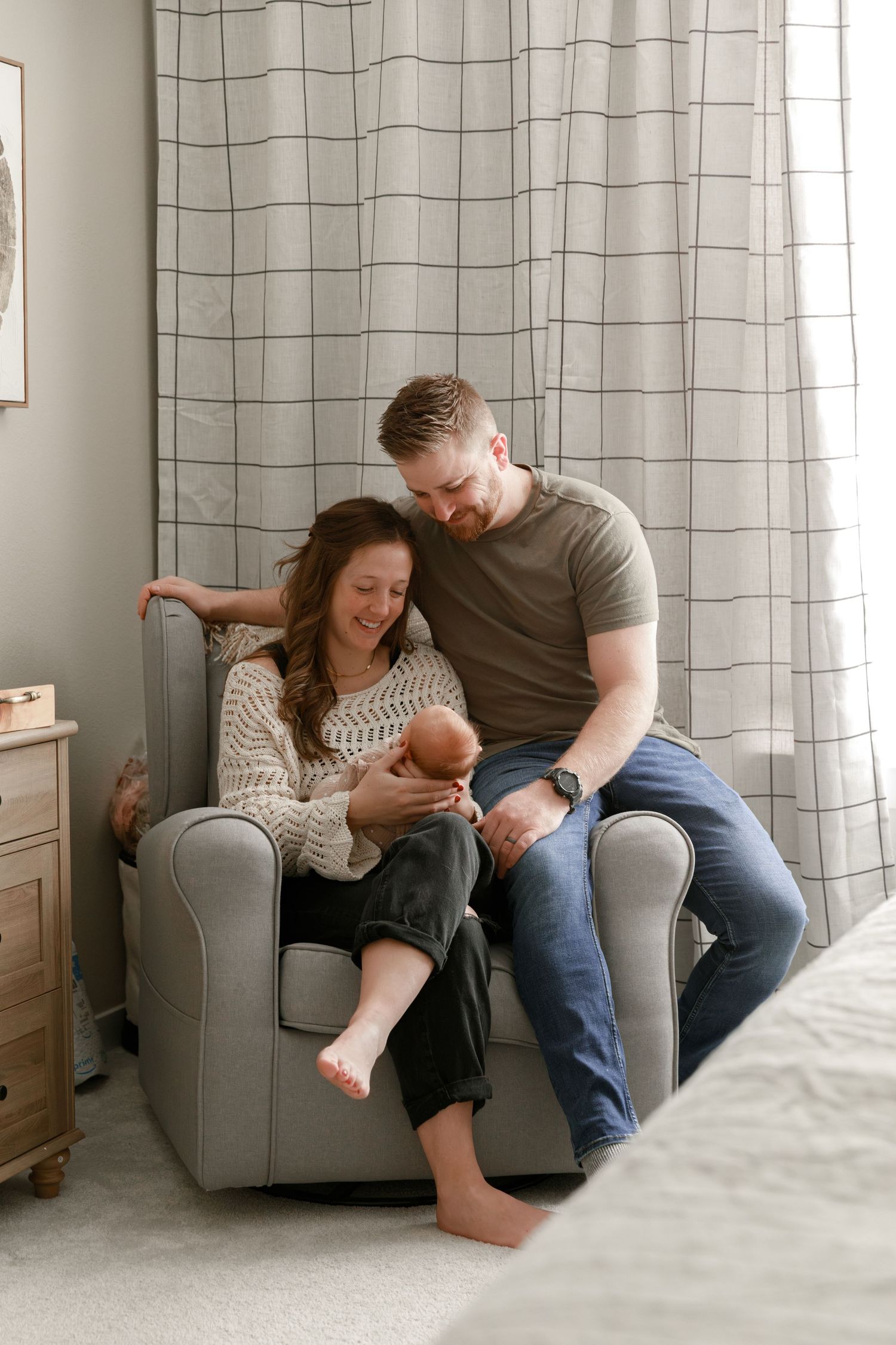 A couple sits together on a grey armchair in a cozy room with plaid curtains.