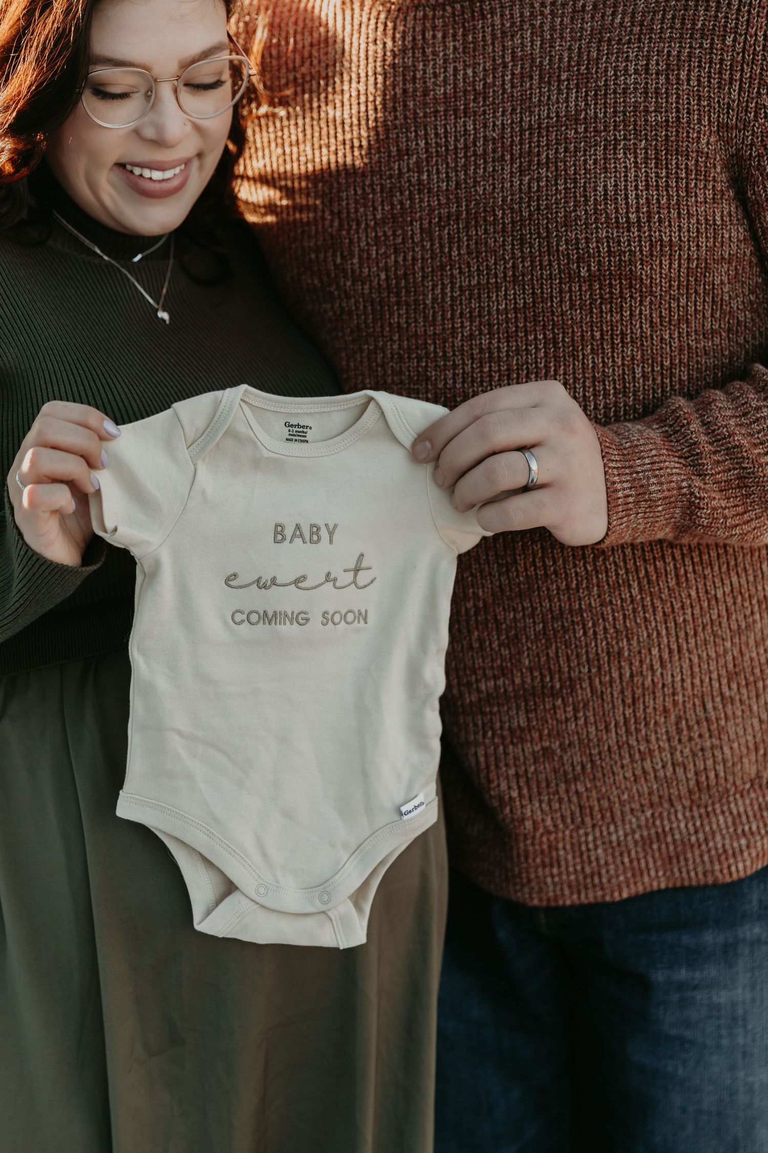 Two people hold up a white baby onesie with text against a brown background.