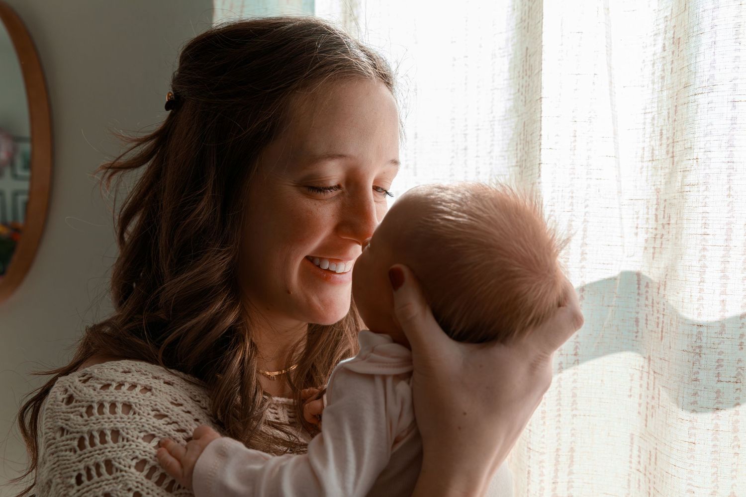An intimate portrait of a mother cradling her newborn baby near a sunlit window.