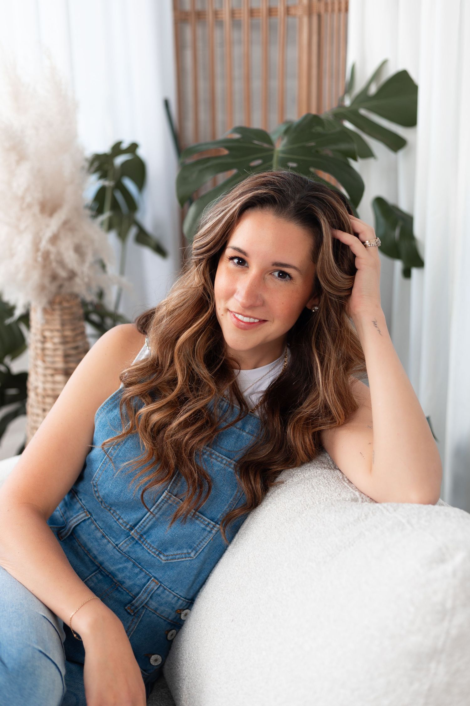 Indoor portrait session with person in denim outfit lounging on white furniture with tropical plants.