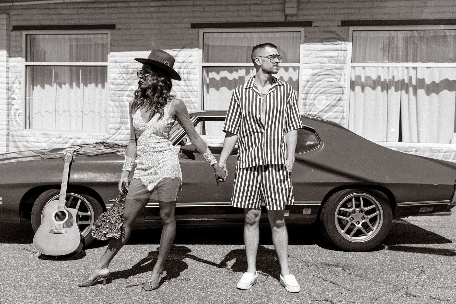 Retro-styled couple posing next to vintage car with guitar in black and white.