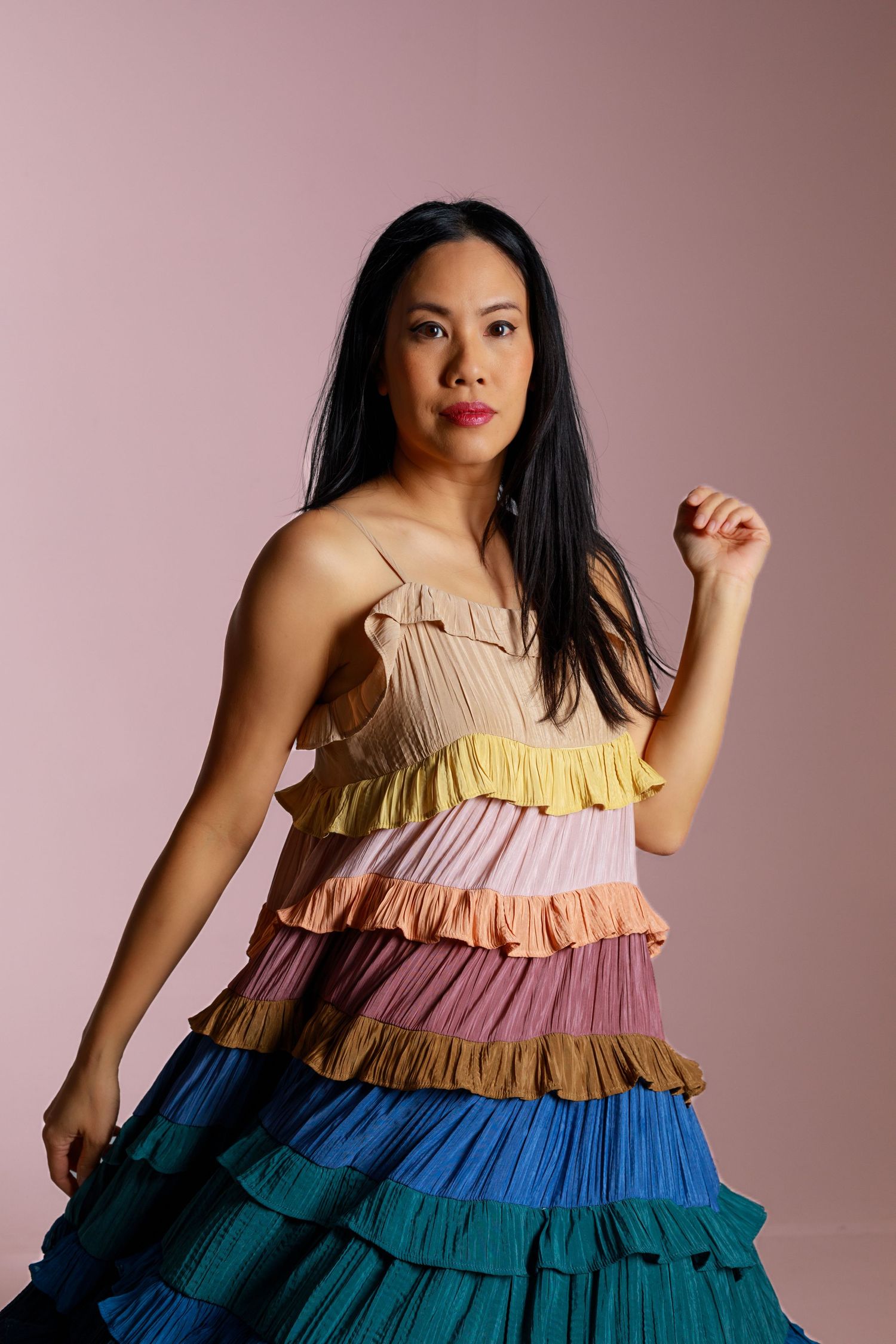 Dynamic fashion shot showcasing multicolored tiered dress with flowing ruffles against rose-colored backdrop.