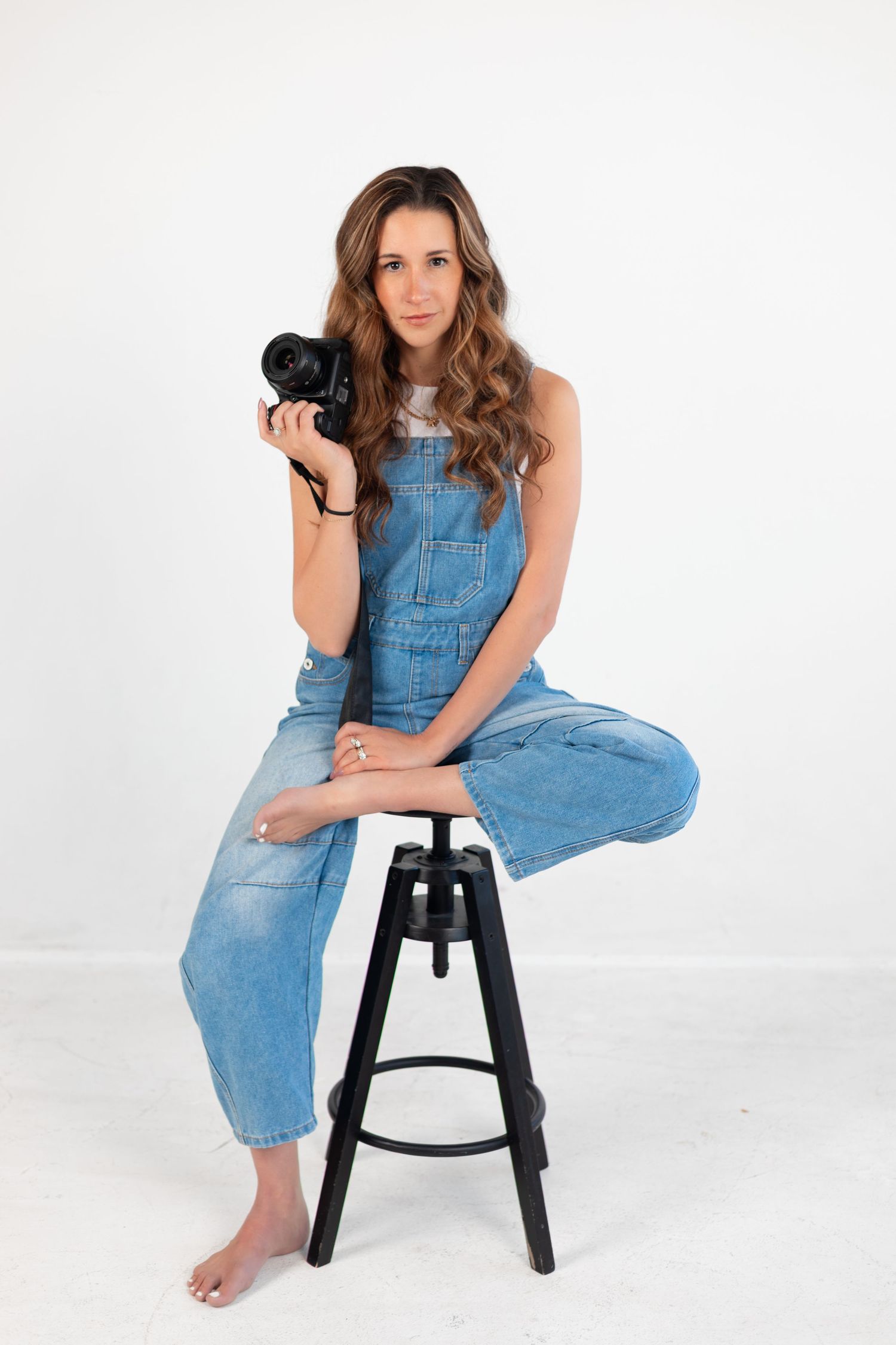 A woman in denim overalls poses casually on a black stool against white backdrop.