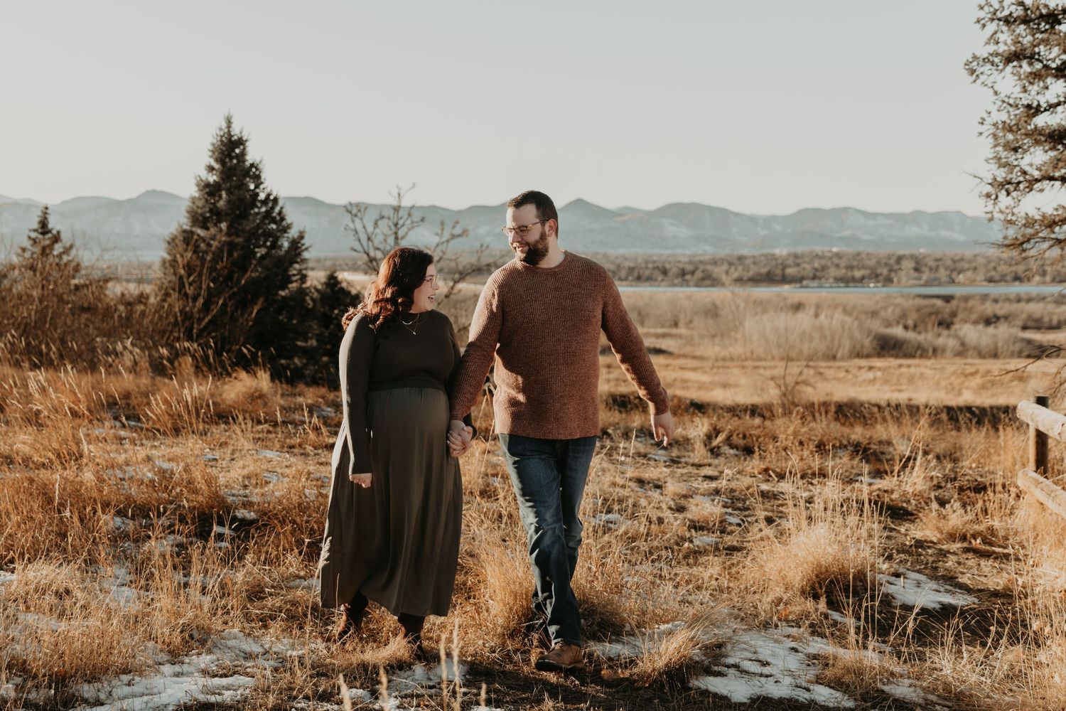 Couple holding hands while walking through golden fields at sunset with mountains in the distance.