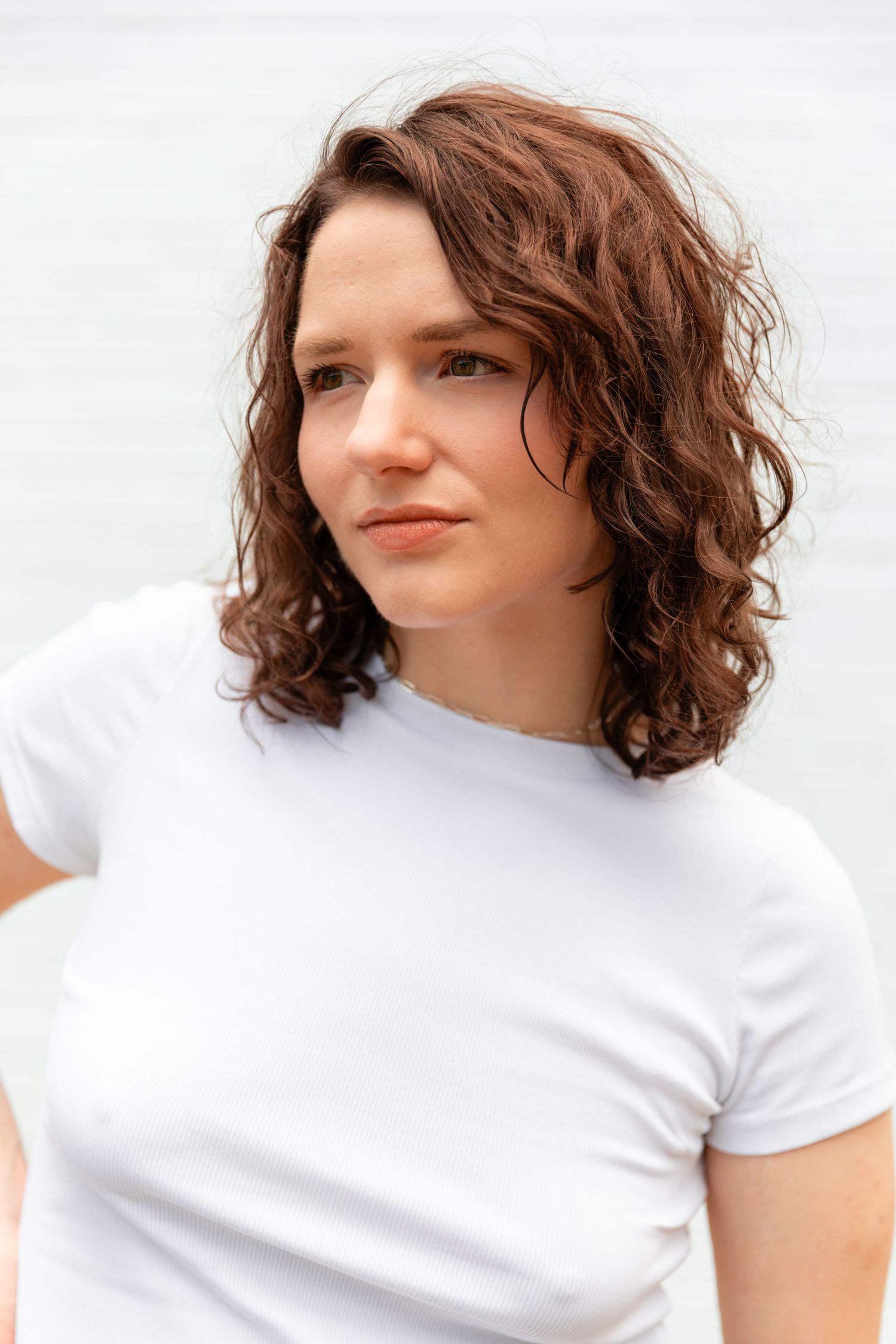 Young woman with curly brown hair wearing a white t-shirt, posing outdoors near water.