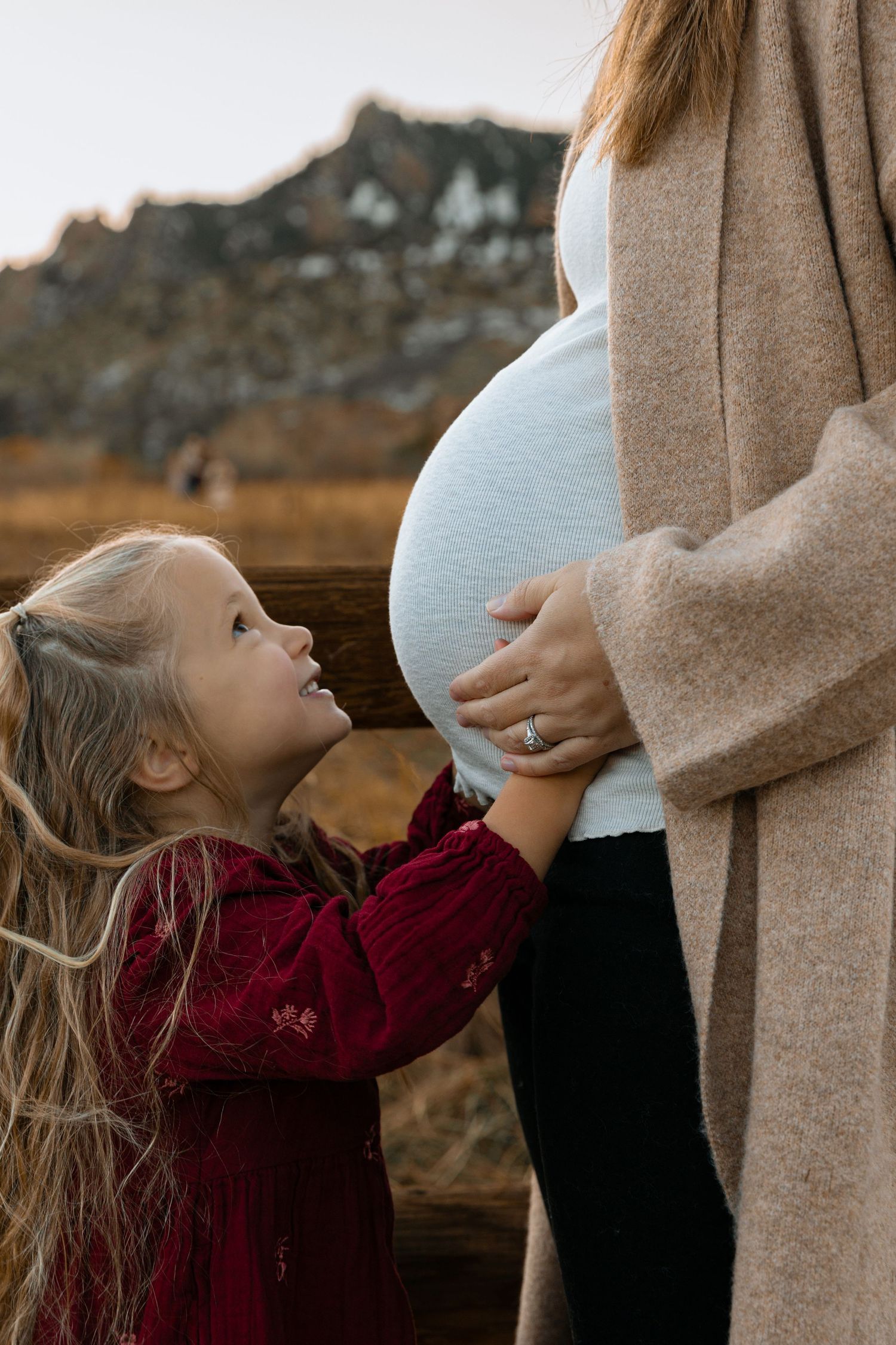 A touching moment between a child in red and an expectant mother against a mountain backdrop.