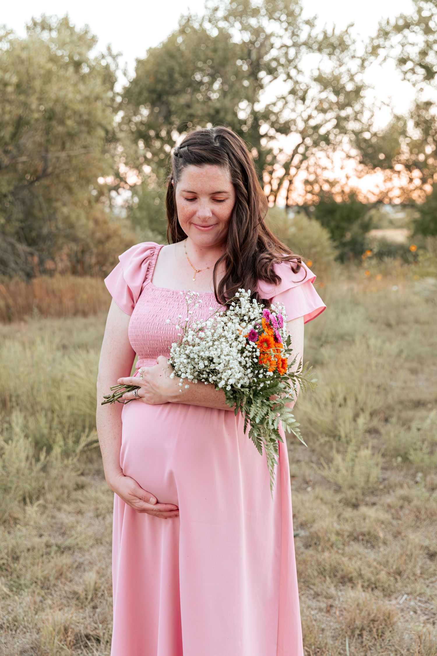 Maternity photos in a field show person in pink dress holding wildflowers and baby's breath bouquet.
