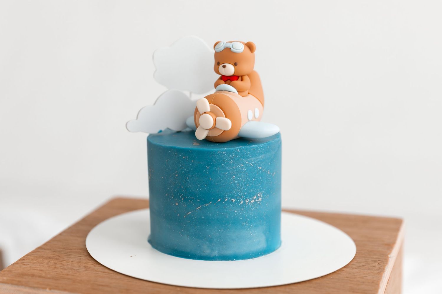 Blue galaxy cake topped with a cute brown teddy bear figurine sitting on white clouds, displayed on a wooden stand.
