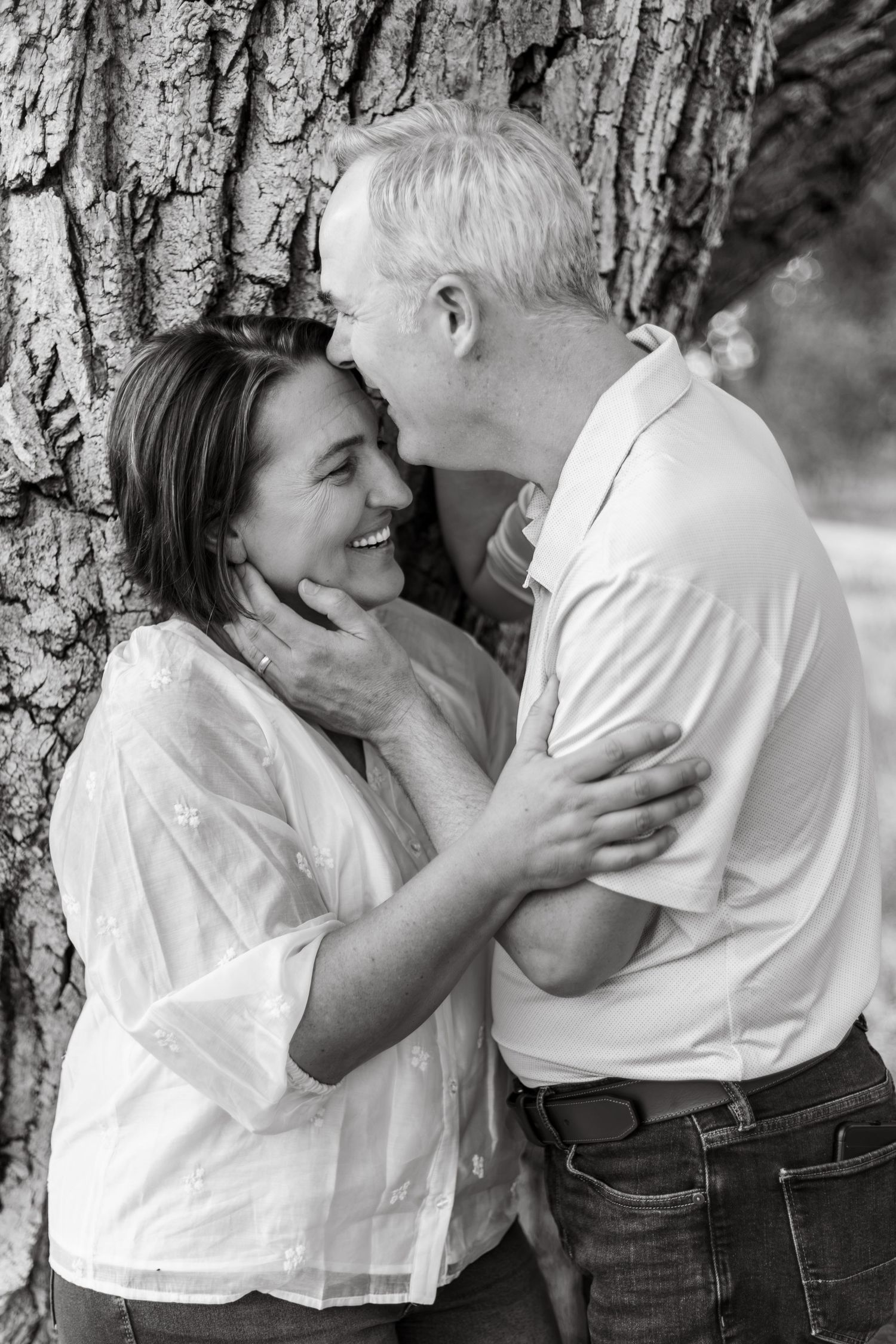 Black and white photo of a smiling couple embracing tenderly against a tree trunk outdoors.